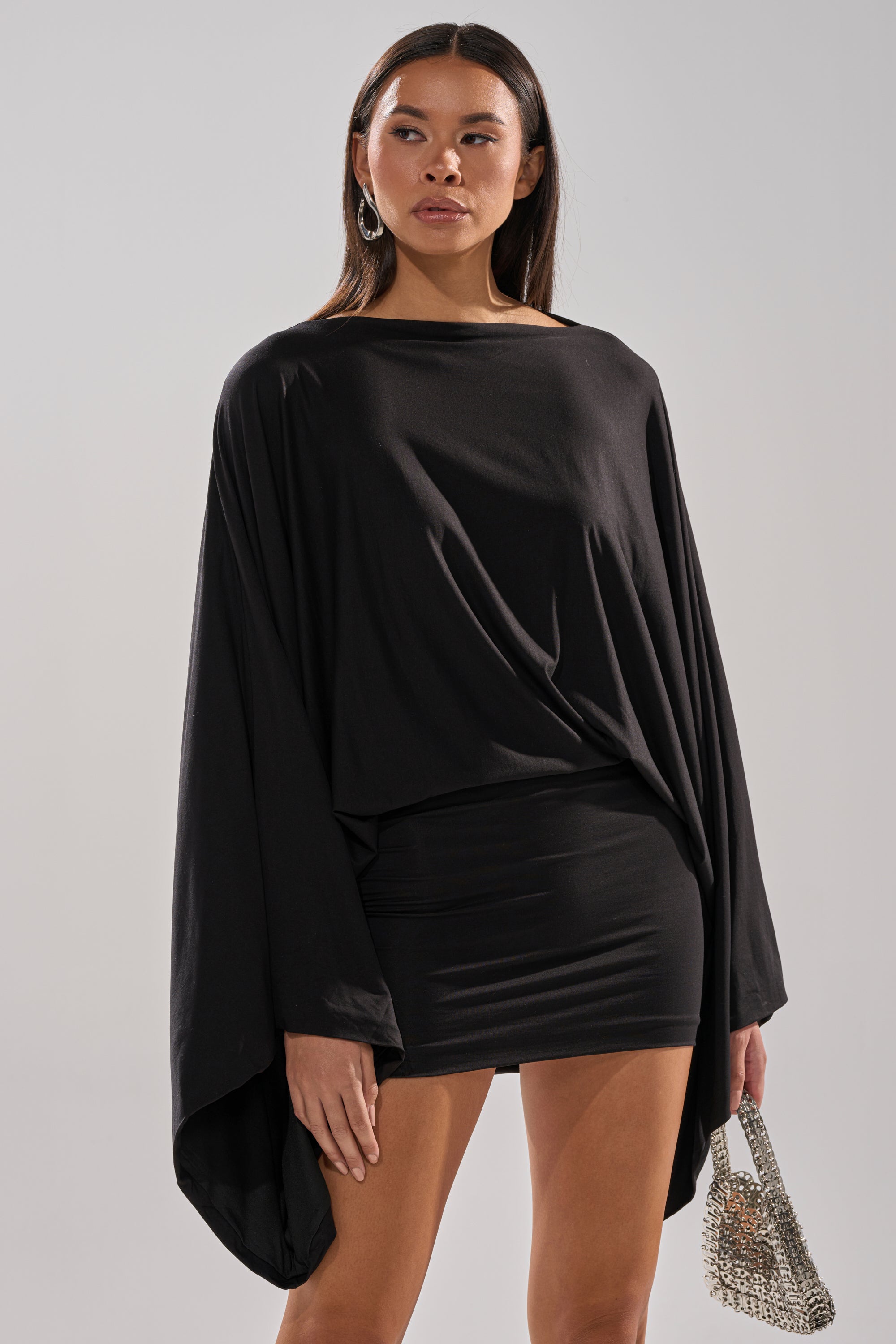 A woman wears the DIANA DRAPED MINI DRESS IN BLACK, featuring batwing sleeves and a fitted mini skirt, paired with a shiny silver purse, posed against a plain light background.