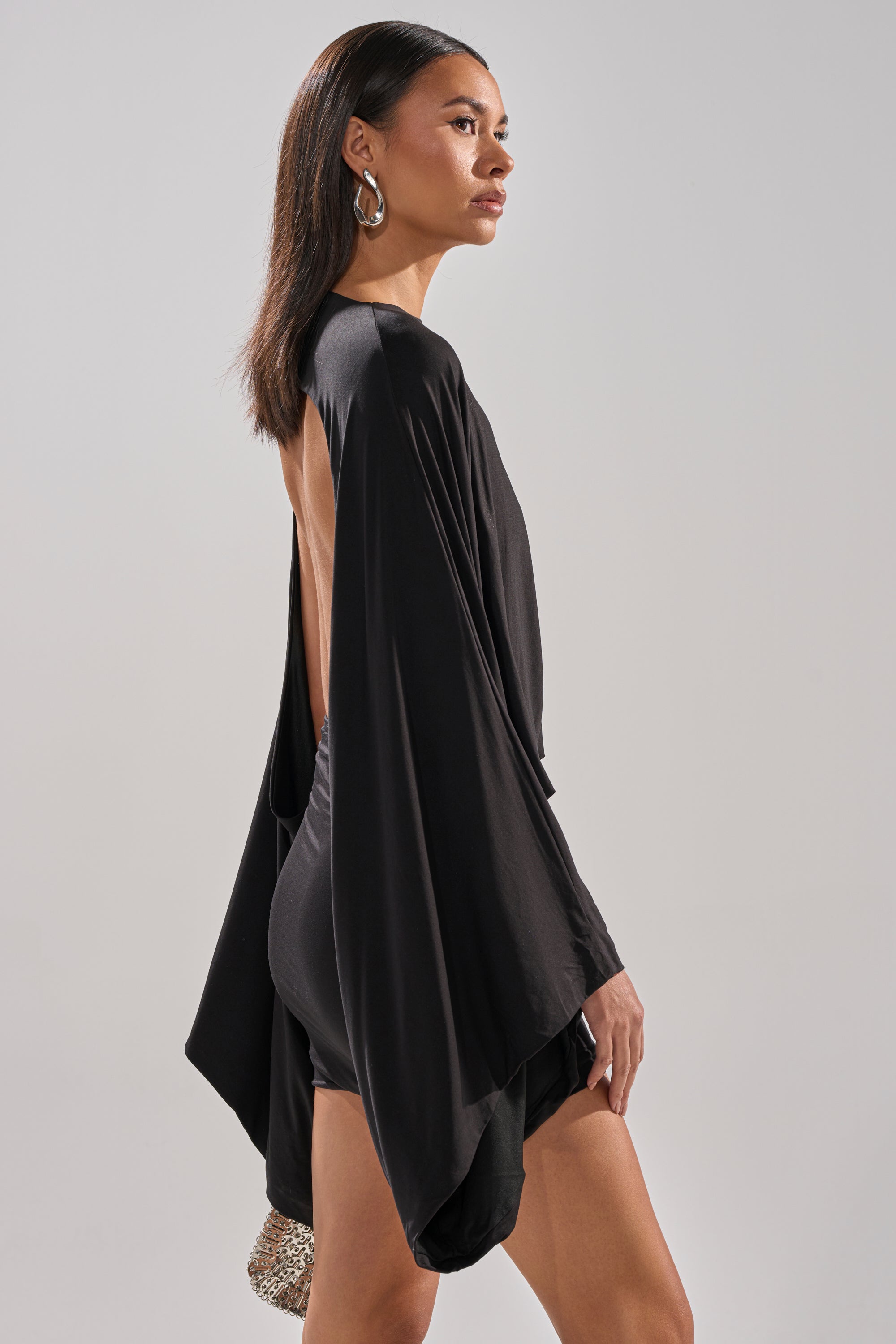 A woman stands in profile against a light gray background, wearing the DIANA DRAPED MINI DRESS IN BLACK with batwing sleeves. She holds a sparkly clutch, has straight dark hair, and accessorizes with large hoop earrings.
