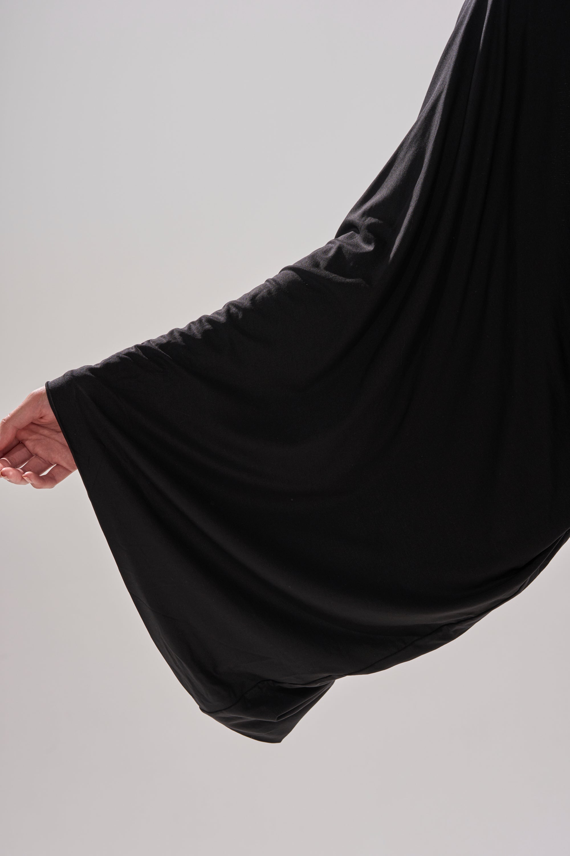A person's left hand extends outward, holding the edge of the loose, flowing sleeve of the DIANA DRAPED MINI DRESS IN BLACK against a plain light background.