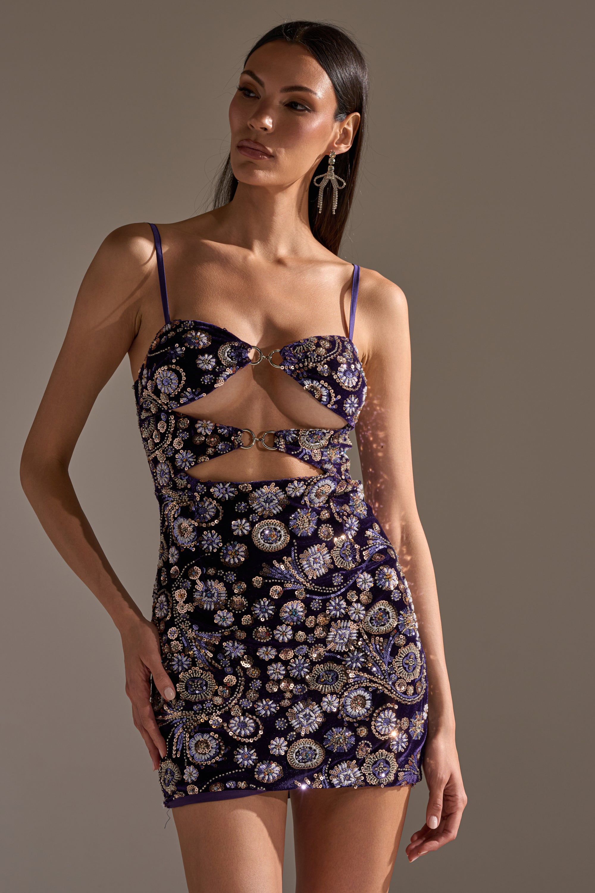 A woman poses in the RUNAWAY BEADED MINI DRESS, a sleeveless dark purple mini dress with gold and silver floral beadwork and front cutouts. She pairs it with statement earrings and styles her long straight hair gracefully.