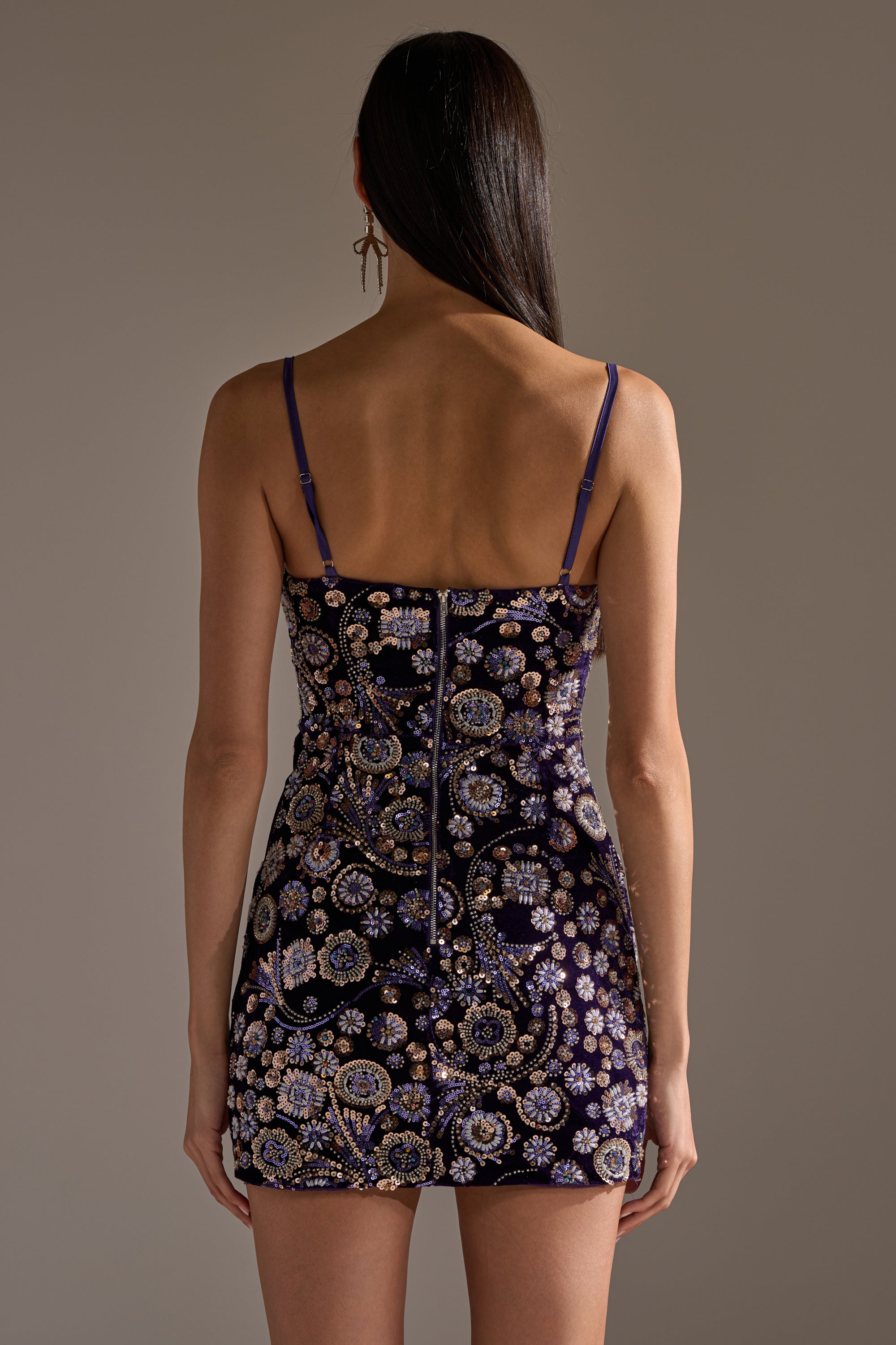 A woman with long dark hair faces away, wearing the RUNAWAY BEADED MINI DRESS— a sleeveless mini dress with thin straps, gold and light blue floral beading, and a visible back zipper for added detail.