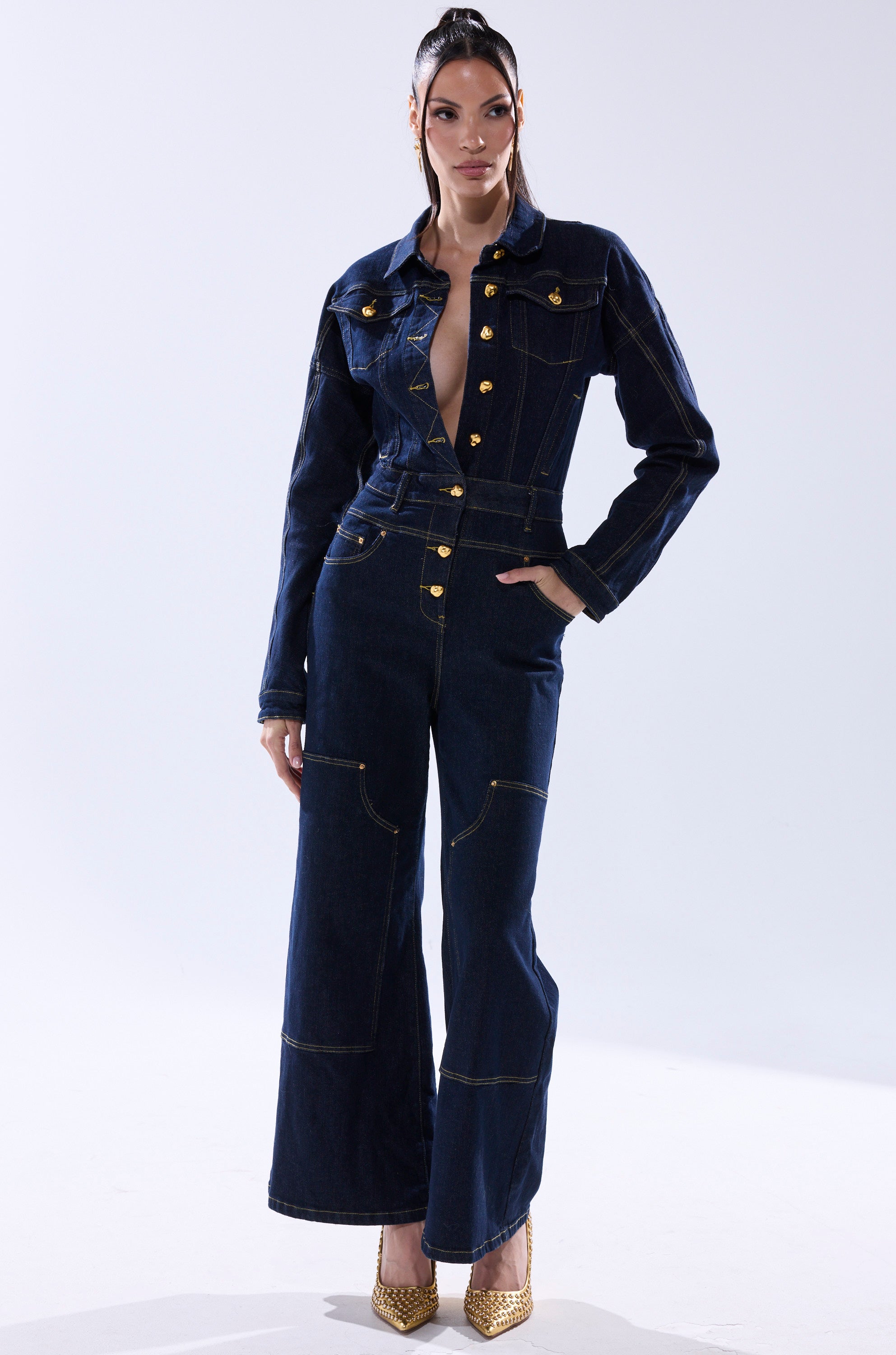 A woman stands confidently against a plain, light background wearing the ANGELA CARGO DENIM JUMPSUIT—a dark blue, long-sleeved utility design with gold buttons, wide legs, and paired with gold heels. 1