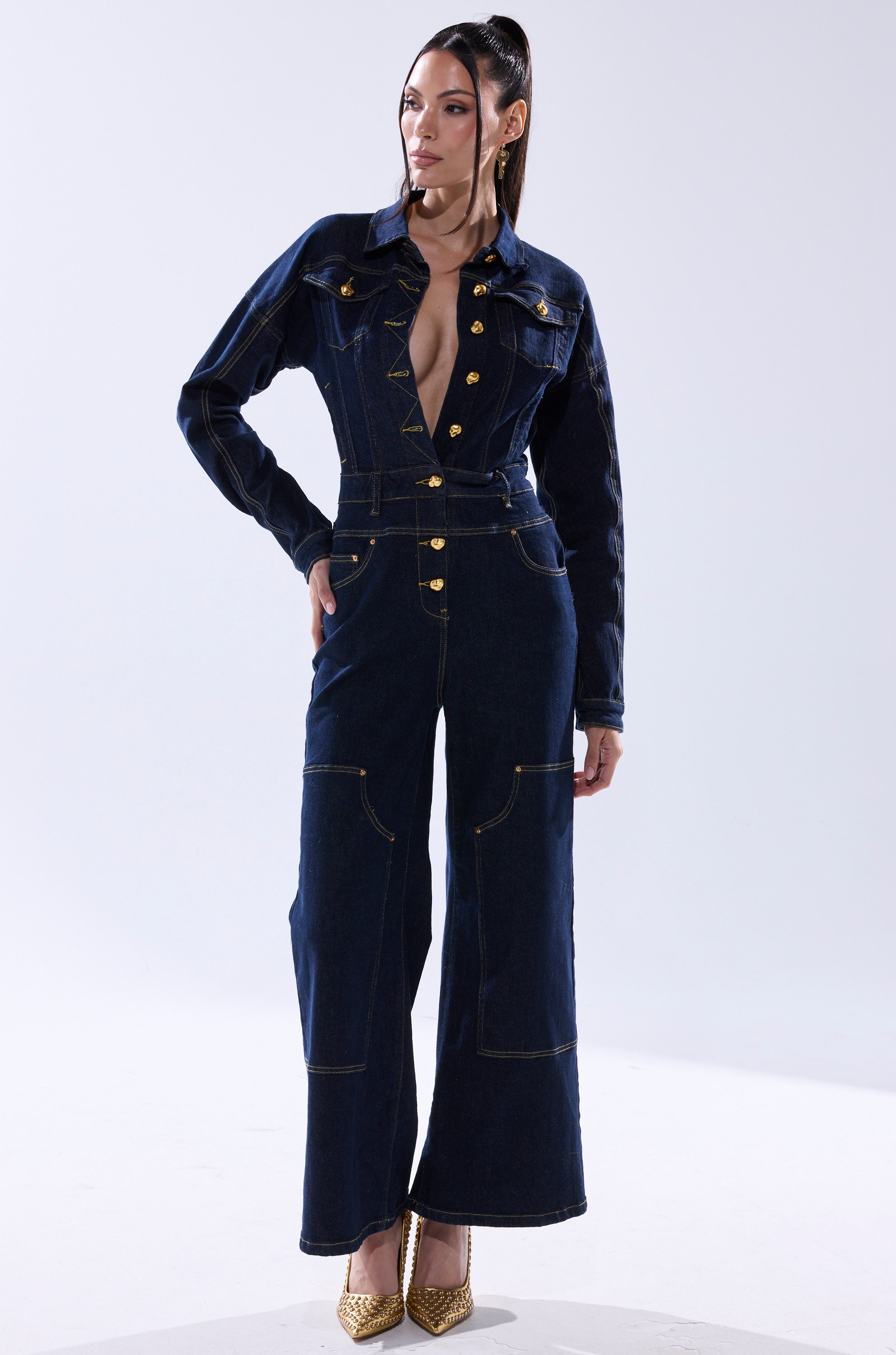A woman stands confidently in the ANGELA CARGO DENIM JUMPSUIT with wide legs and gold buttons, her hair in a sleek ponytail. She completes the look with gold high-heeled shoes against a plain, light-colored background. 2