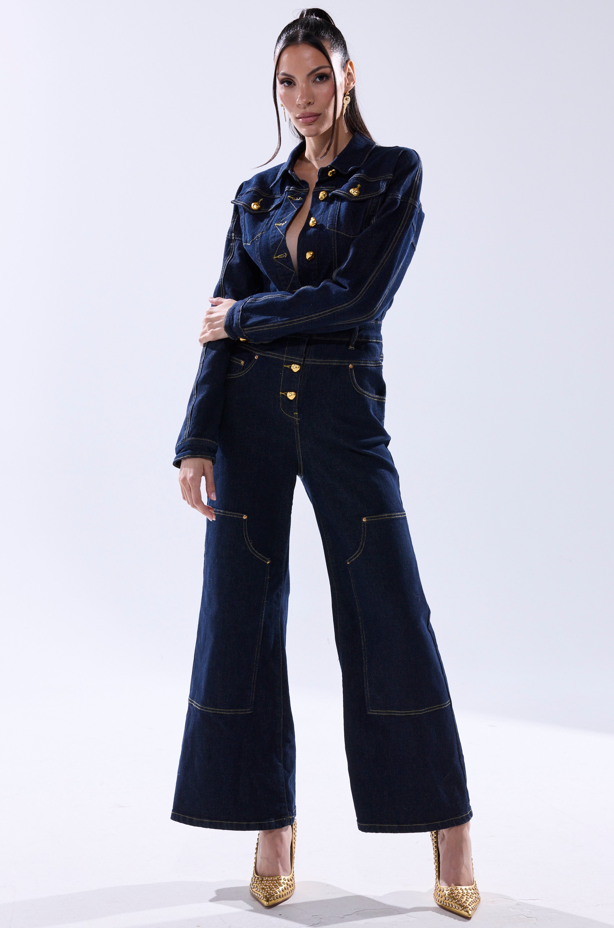 A woman stands confidently in the ANGELA CARGO DENIM JUMPSUIT, featuring gold buttons and wide legs, paired with gold high heels against a plain light background. 3