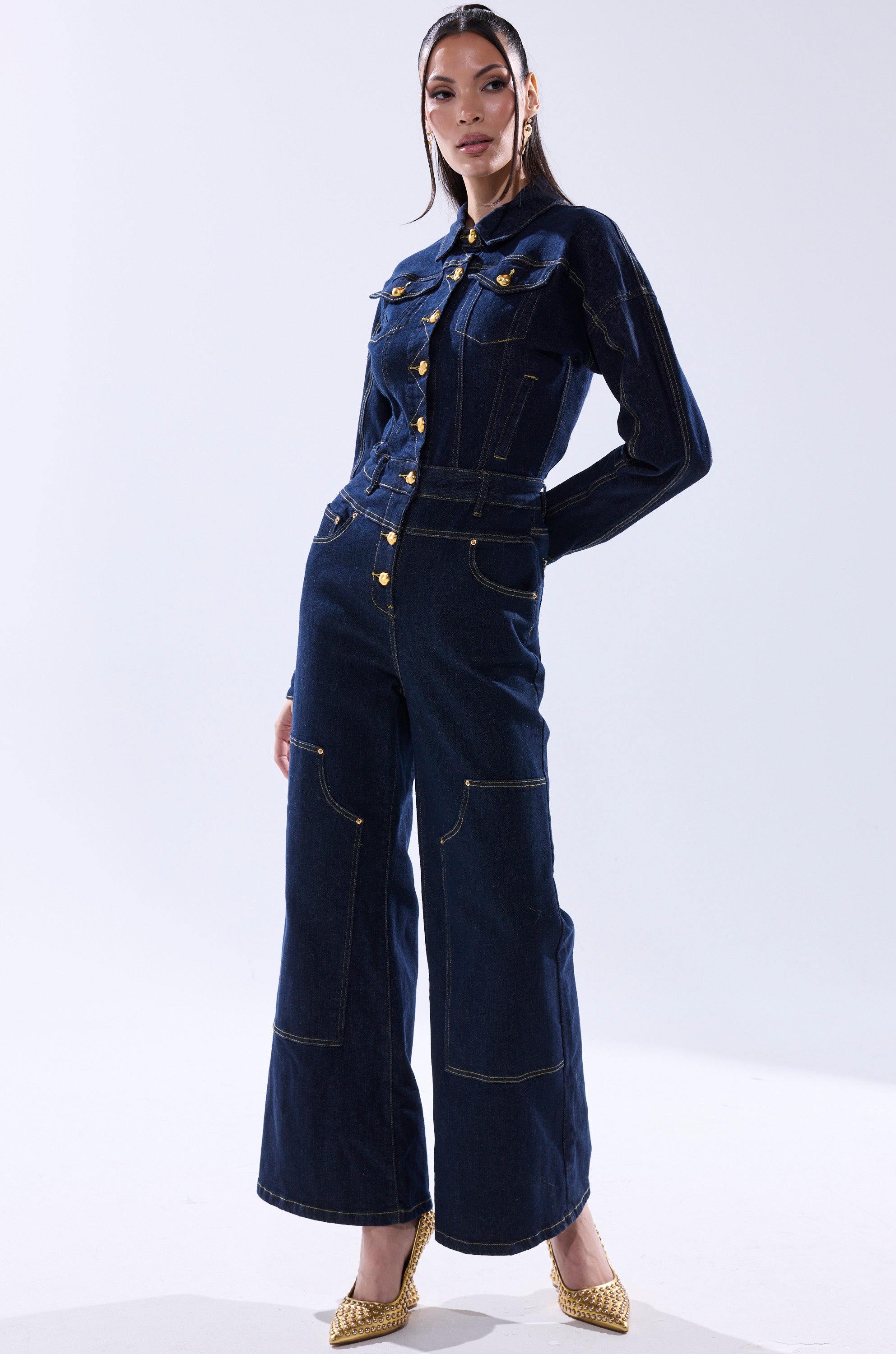 A woman with straight hair in a headband wears the ANGELA CARGO DENIM JUMPSUIT, featuring gold buttons and stitching, paired with pointed gold heels, standing against a plain background. 4