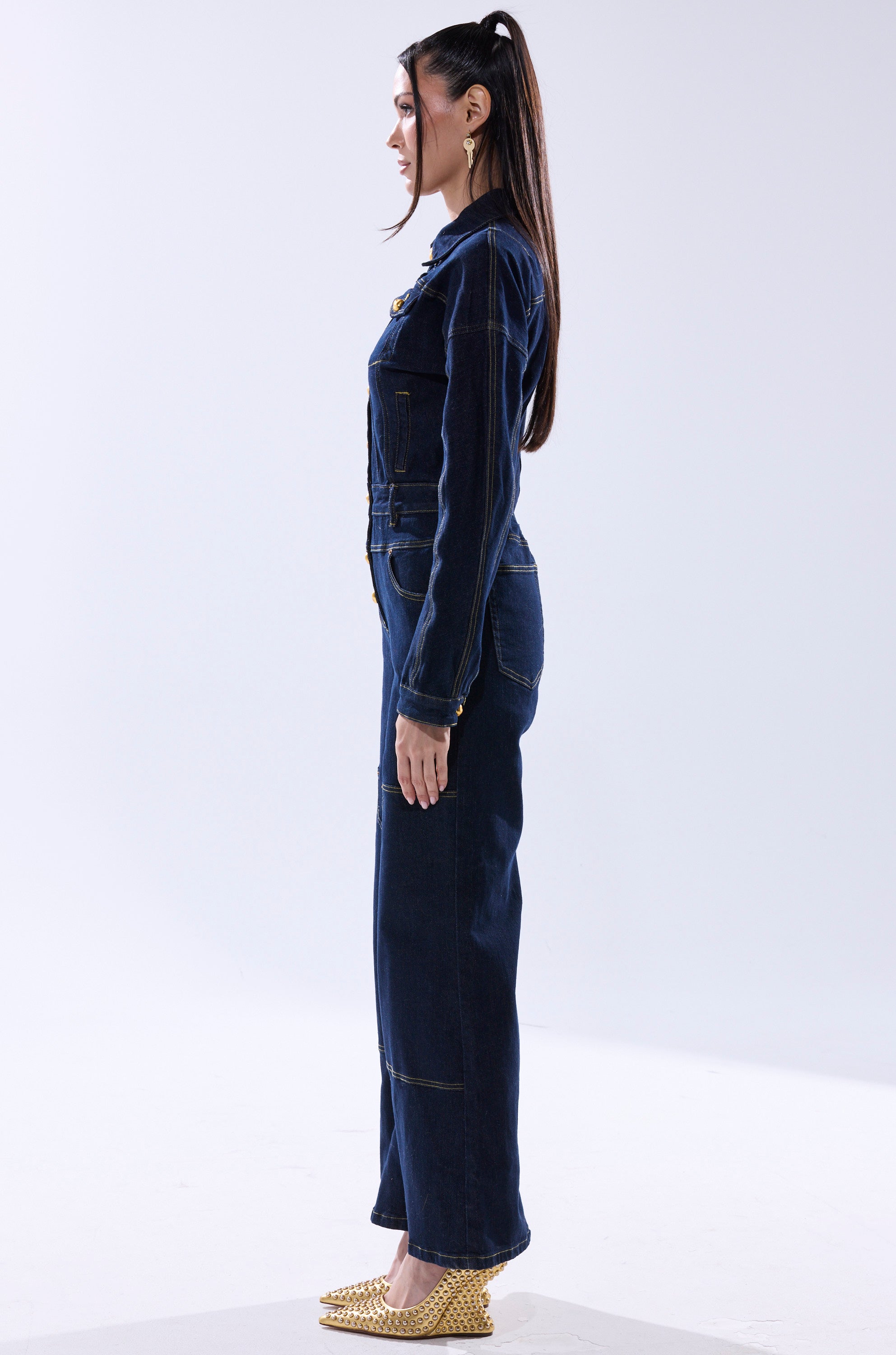 A woman with long straight hair stands in profile, wearing the chic ANGELA CARGO DENIM JUMPSUIT and gold pointed shoes against a plain light background. 5