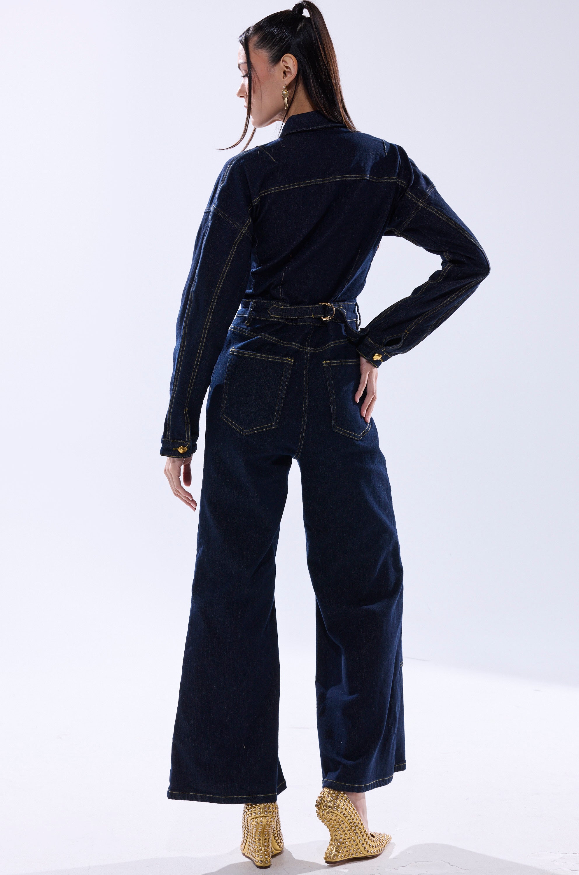 A woman with a high ponytail stands against a plain white background, wearing the ANGELA CARGO DENIM JUMPSUIT with long sleeves and wide-leg pants, paired with gold textured heeled ankle boots. 6