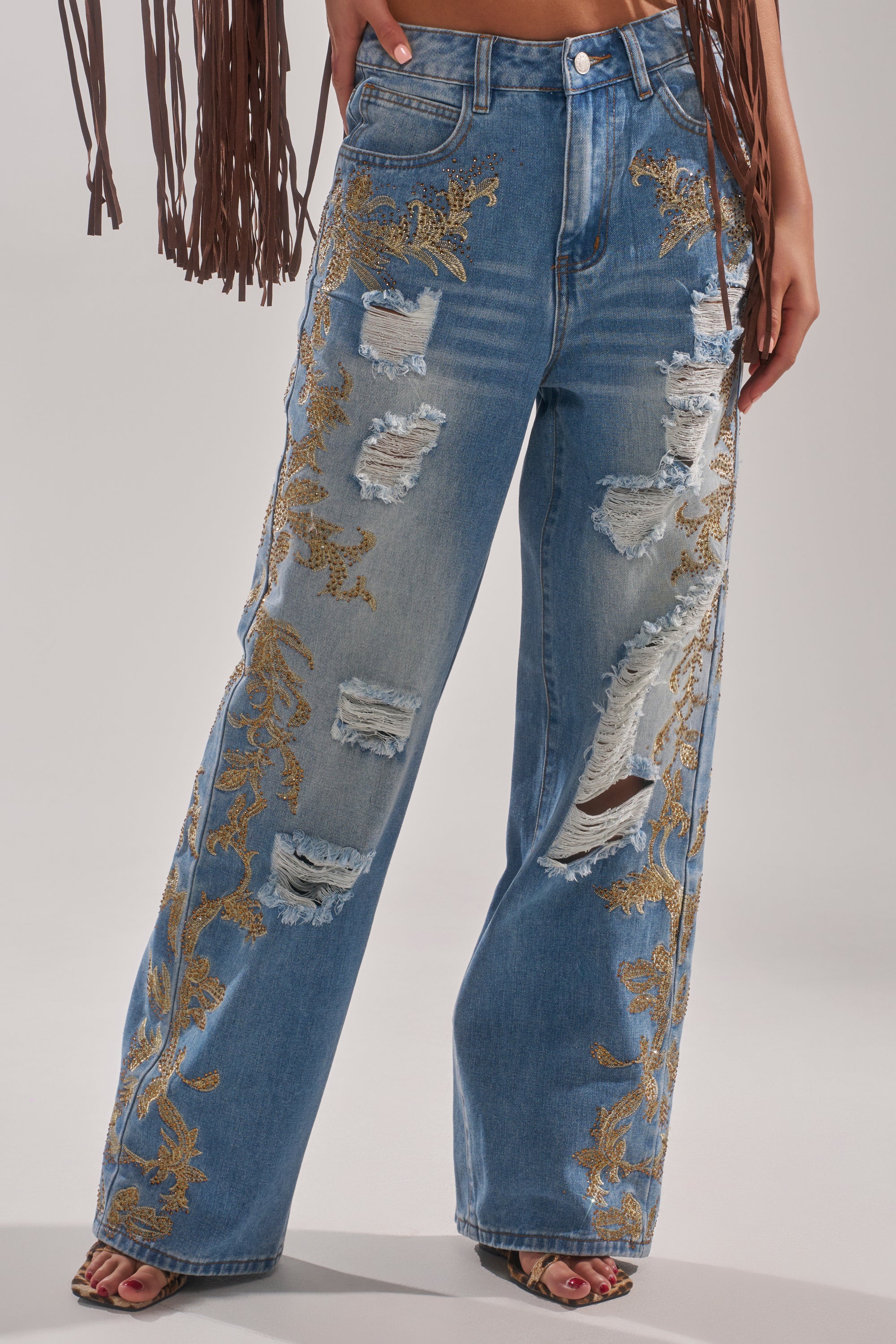 A person wears the CLEO GOLDEN DISTRESSED STRAIGHT DENIM PANT—high-waisted blue jeans with gold embroidery, distressed rips, and wide legs—paired with heeled sandals. Brown fringe drapes from the top left. 0