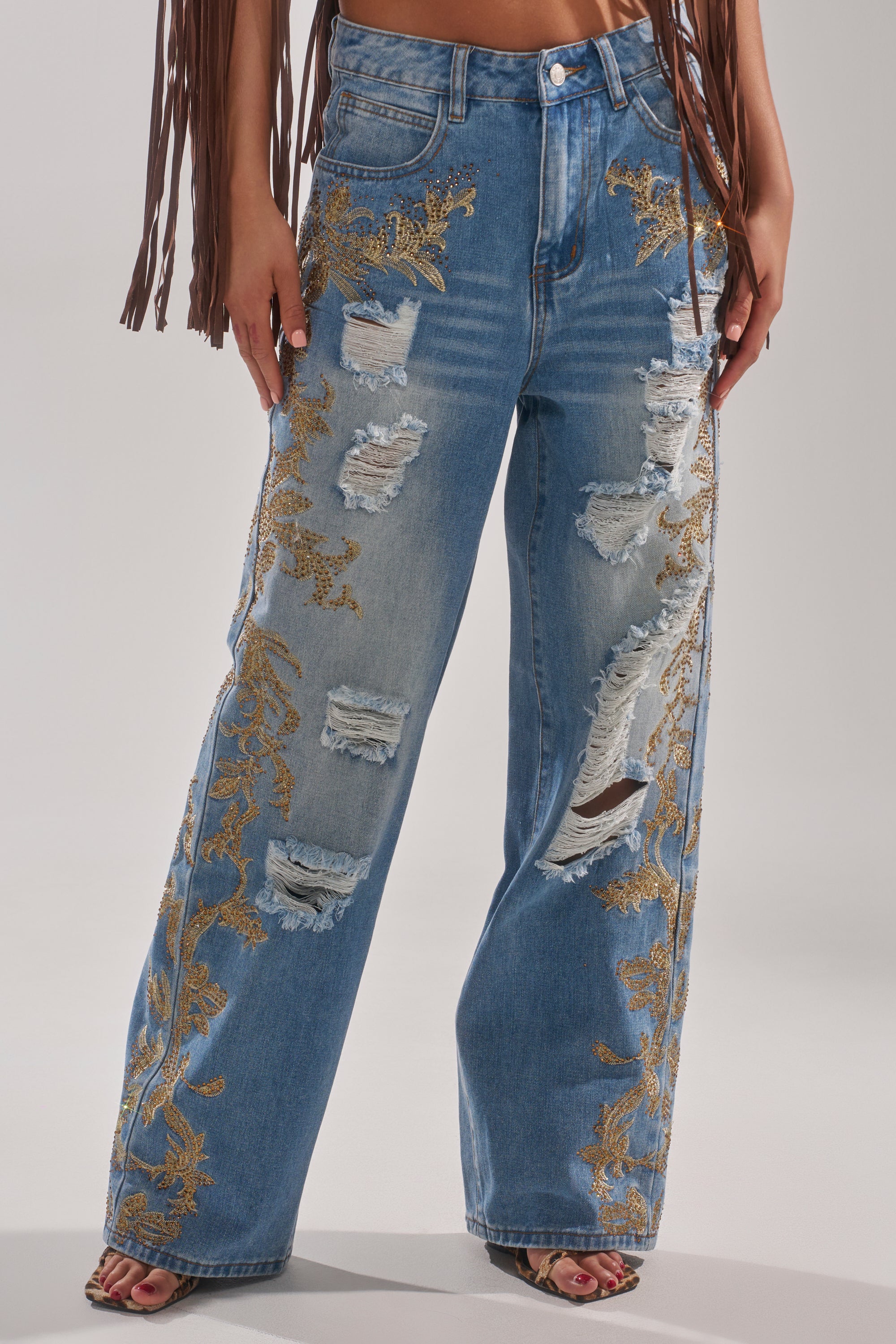 Wearing CLEO GOLDEN DISTRESSED STRAIGHT DENIM PANT with ripped details and gold embroidery, paired with open-toe shoes. Brown fringe from a top or jacket adds texture near the arms to complement the embroidered patches. 1