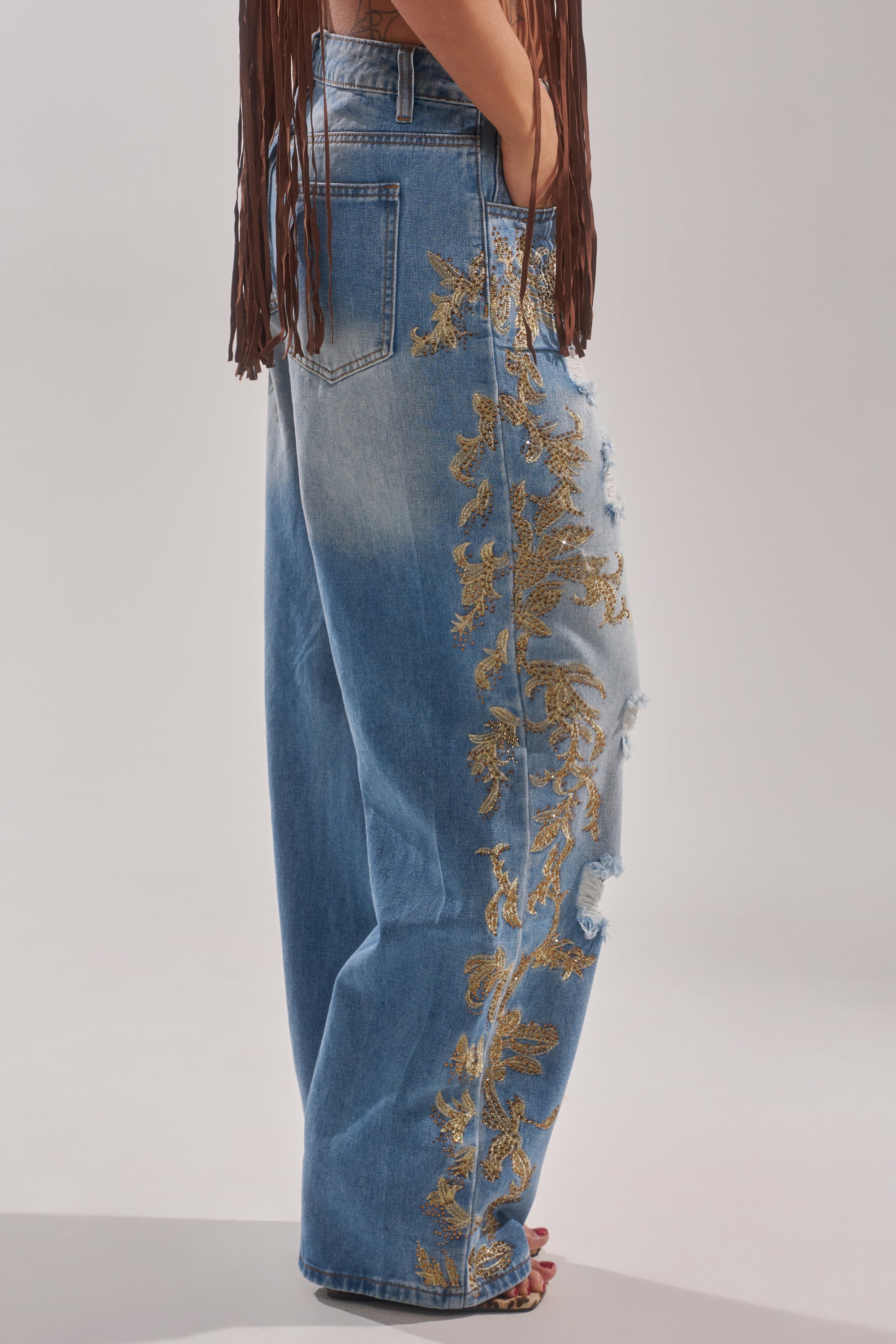 A person wearing CLEO GOLDEN DISTRESSED STRAIGHT DENIM PANT featuring loose fit, gold floral embroidery, and distressed side patches, paired with a brown fringed top. Side waist-down view shown. 2
