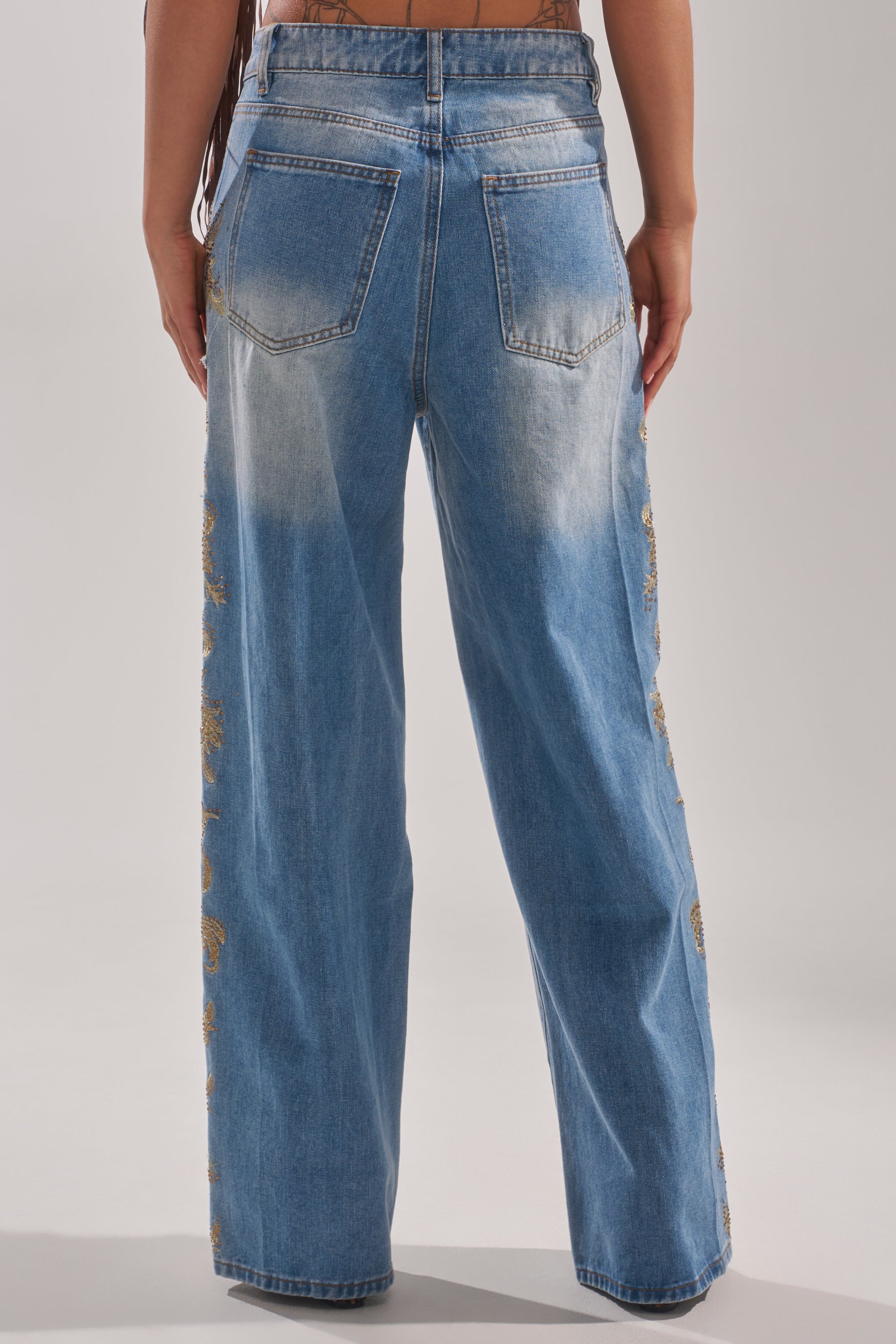 A person is seen from the back in CLEO GOLDEN DISTRESSED STRAIGHT DENIM PANT, a high-waisted, loose-fit blue jean with a faded wash, two back pockets, and gold embroidery along the outer seams. 3