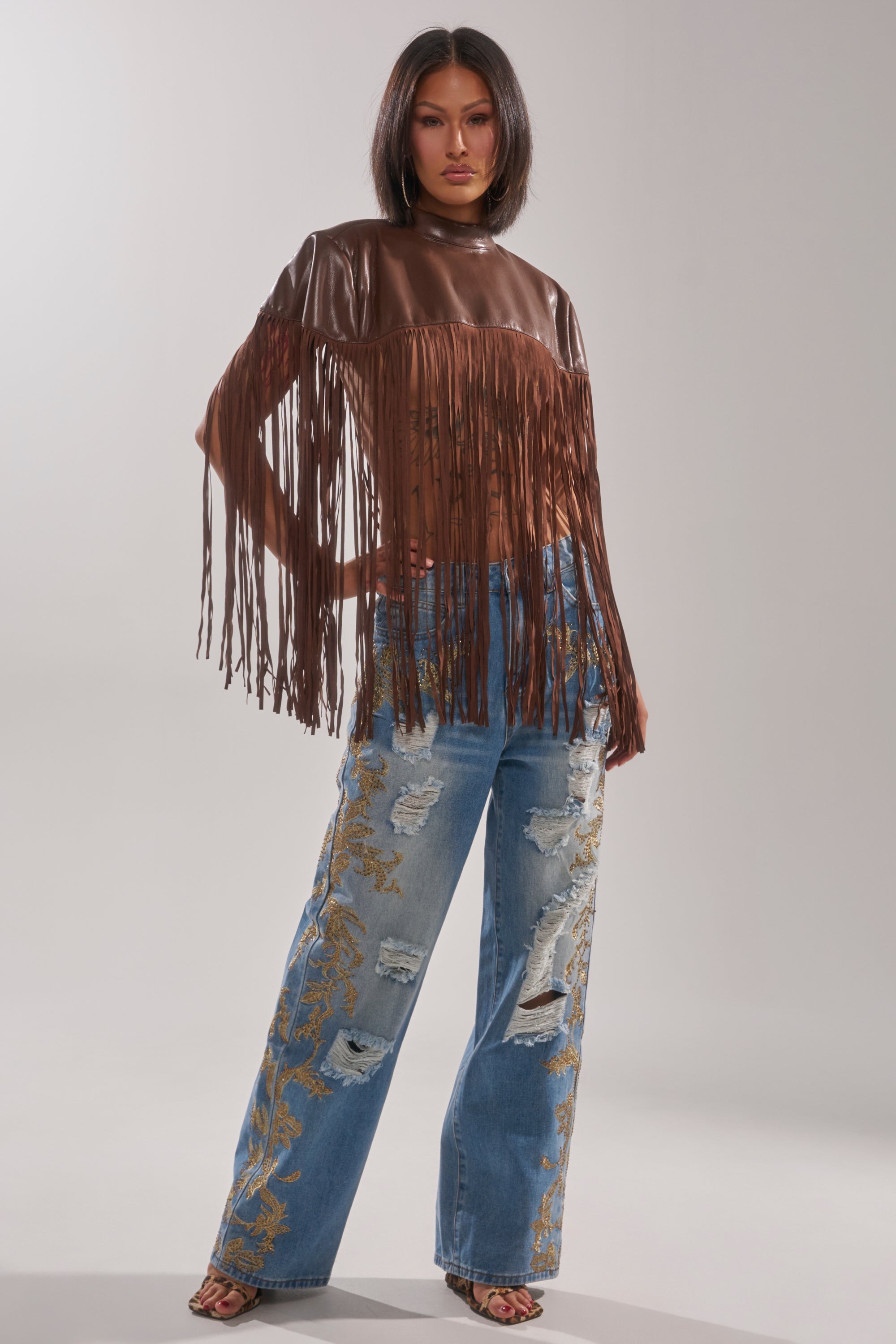 A woman stands confidently in CLEO GOLDEN DISTRESSED STRAIGHT DENIM PANT, paired with a sheer brown fringed top. Her straight dark hair frames her look, completed with open-toe shoes and chic golden embroidered denim. 5