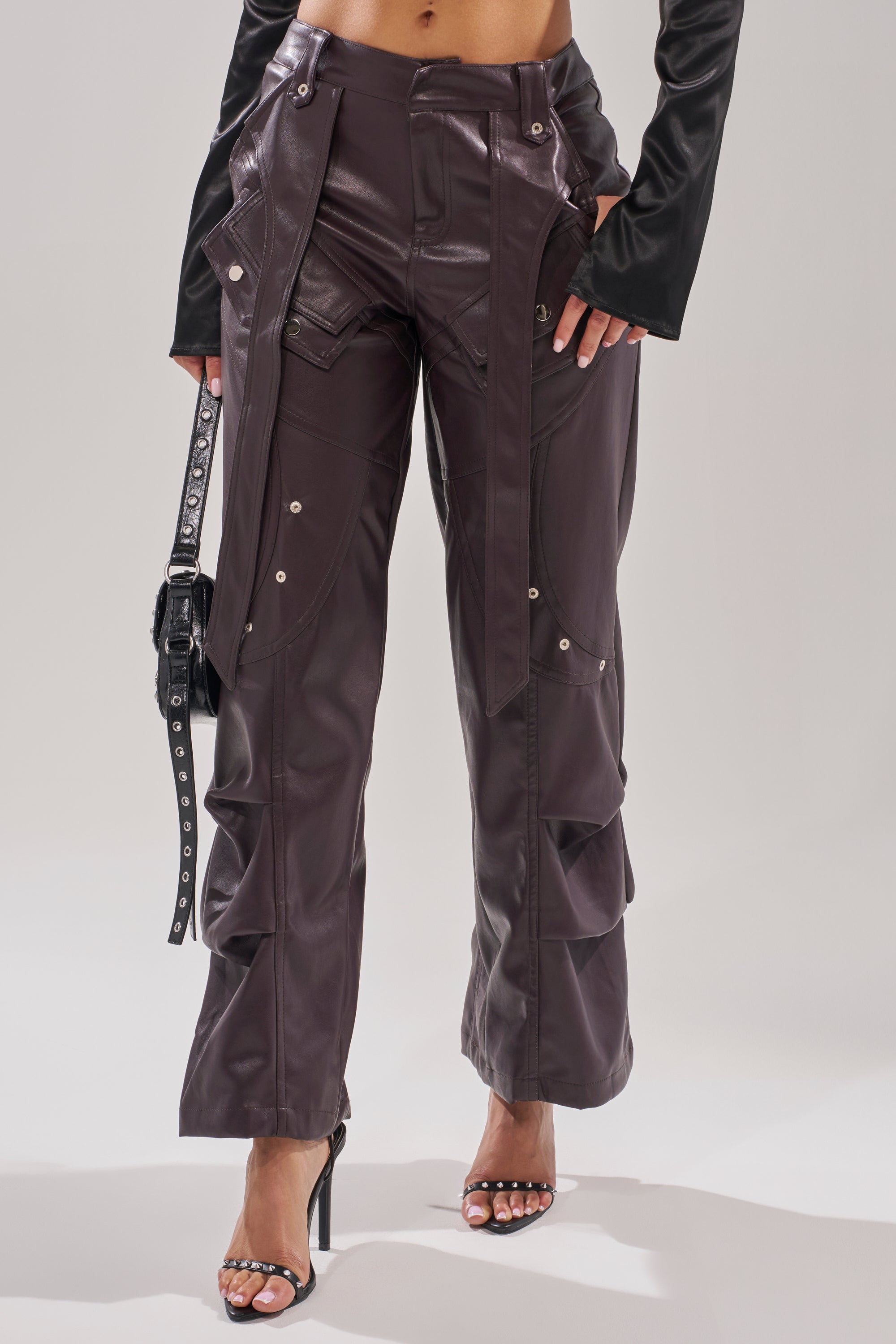 Wearing THE BEST PLUM FAUX LEATHER CARGO TROUSER—high-waisted, dark brown, wide-leg pants with silver snaps and straps—paired with a black crop top, black purse, and black heeled sandals. 1