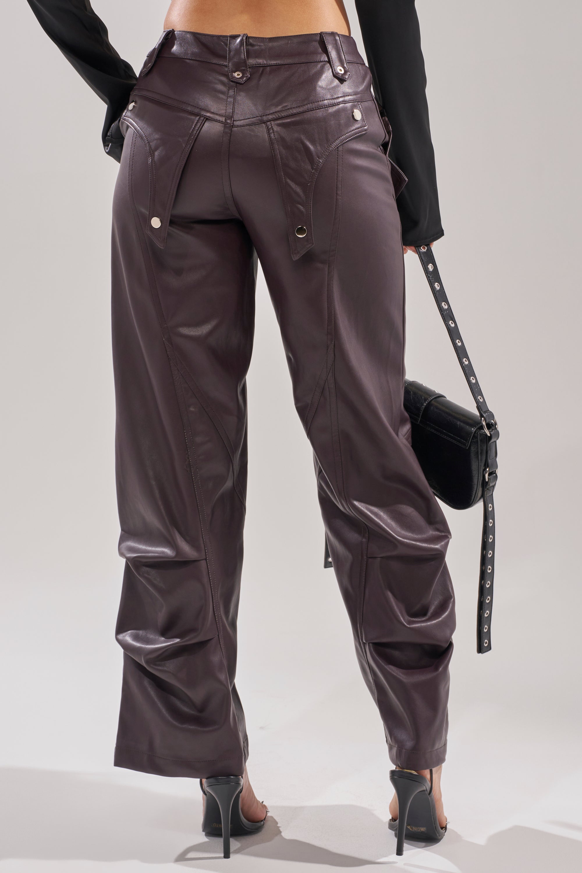 A person faces away, wearing THE BEST PLUM FAUX LEATHER CARGO TROUSER with a wide-leg silhouette and visible seams, paired with high-heeled sandals and carrying a black studded handbag. 3