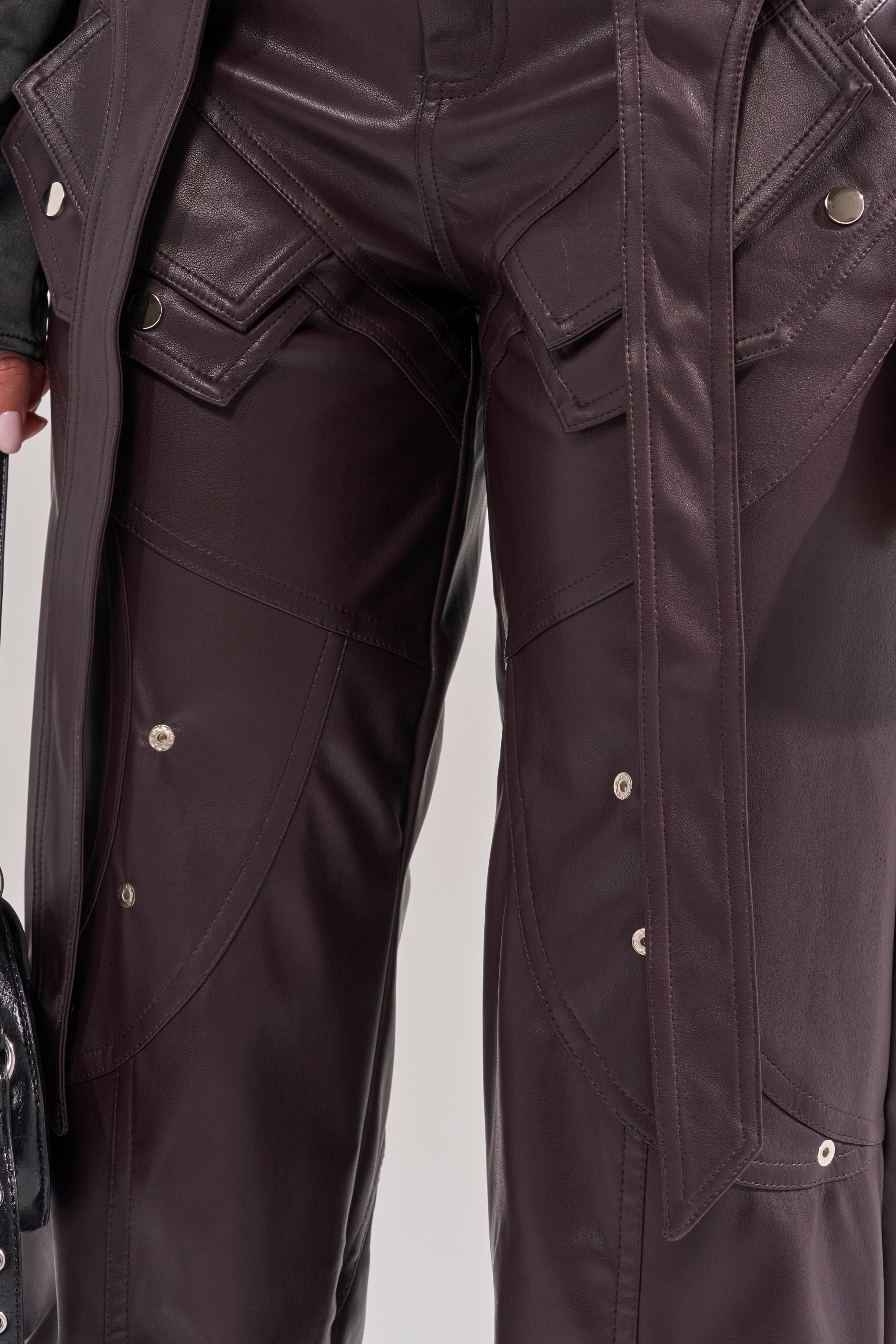 Close-up of a person wearing THE BEST PLUM FAUX LEATHER CARGO TROUSER—high-rise, wide-leg, with decorative stitching, flap pockets, and silver buttons. A matching belted jacket and a black handbag are also shown. 6