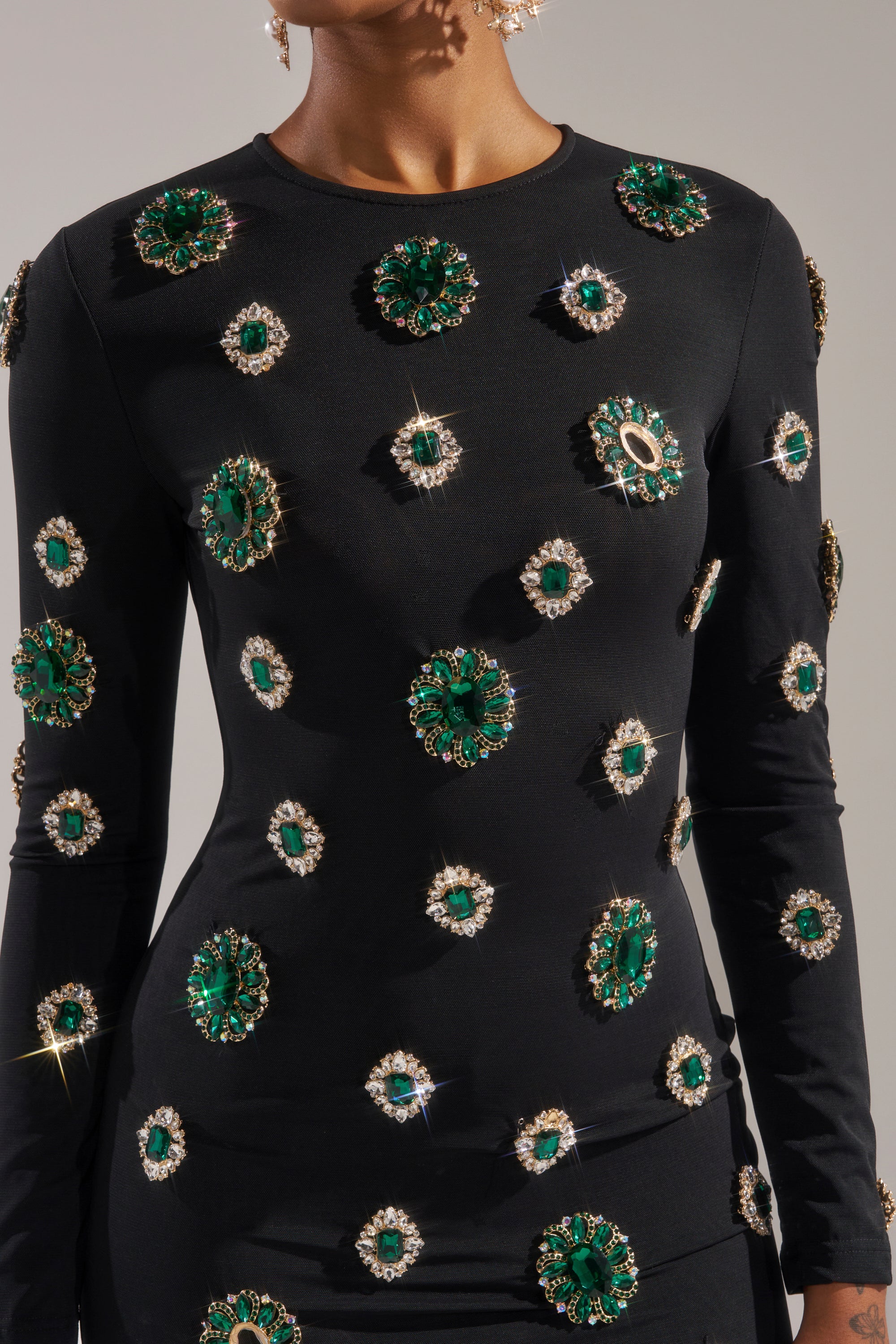 A person in the BLING BLING MESH MINI DRESS WITH RHINESTONE EMBELLISHMENTS, a curve-hugging black long-sleeve dress featuring large green jeweled details, rhinestones, and sparkling crystals. The head is partially out of frame, gems glinting in the light.