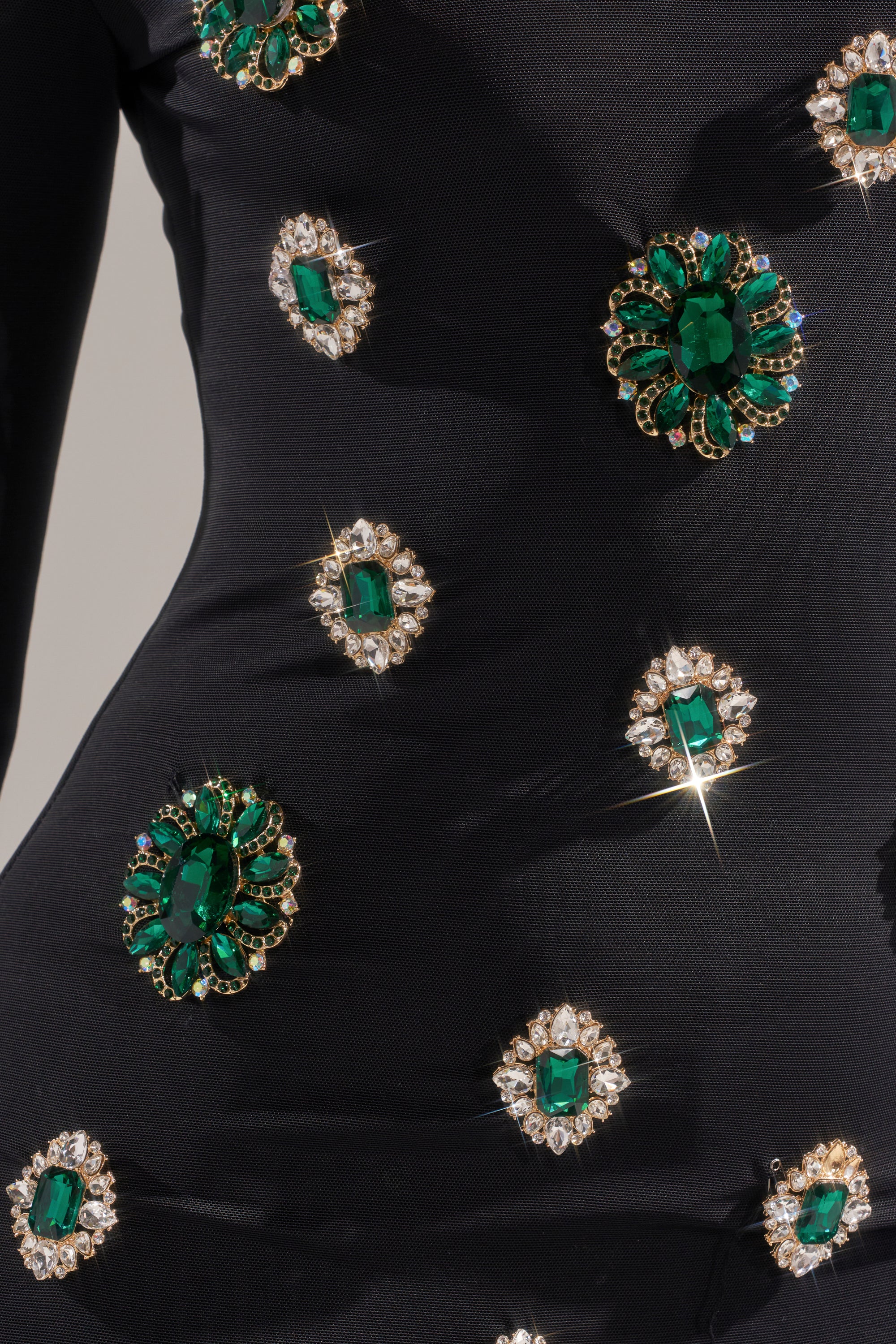 The BLING BLING MESH MINI DRESS WITH RHINESTONE EMBELLISHMENTS features a curve-hugging black silhouette, decorated with sparkling green gemstones and clear crystals in eye-catching patterns.