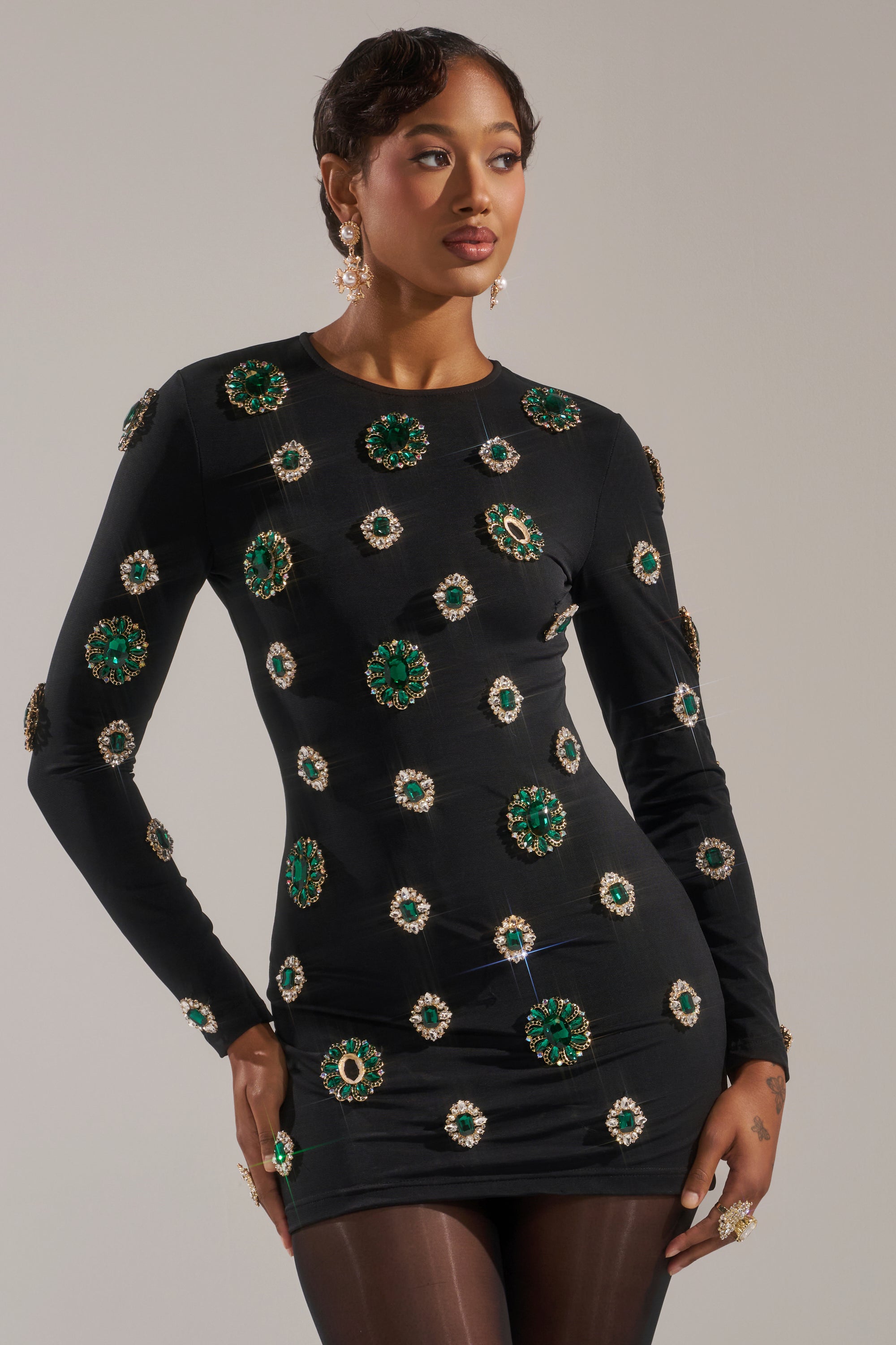 A woman models the BLING BLING MESH MINI DRESS WITH RHINESTONE EMBELLISHMENTS, featuring large green and gold jewels. She pairs it with statement earrings and rings, her hair styled neatly, standing against a plain background.