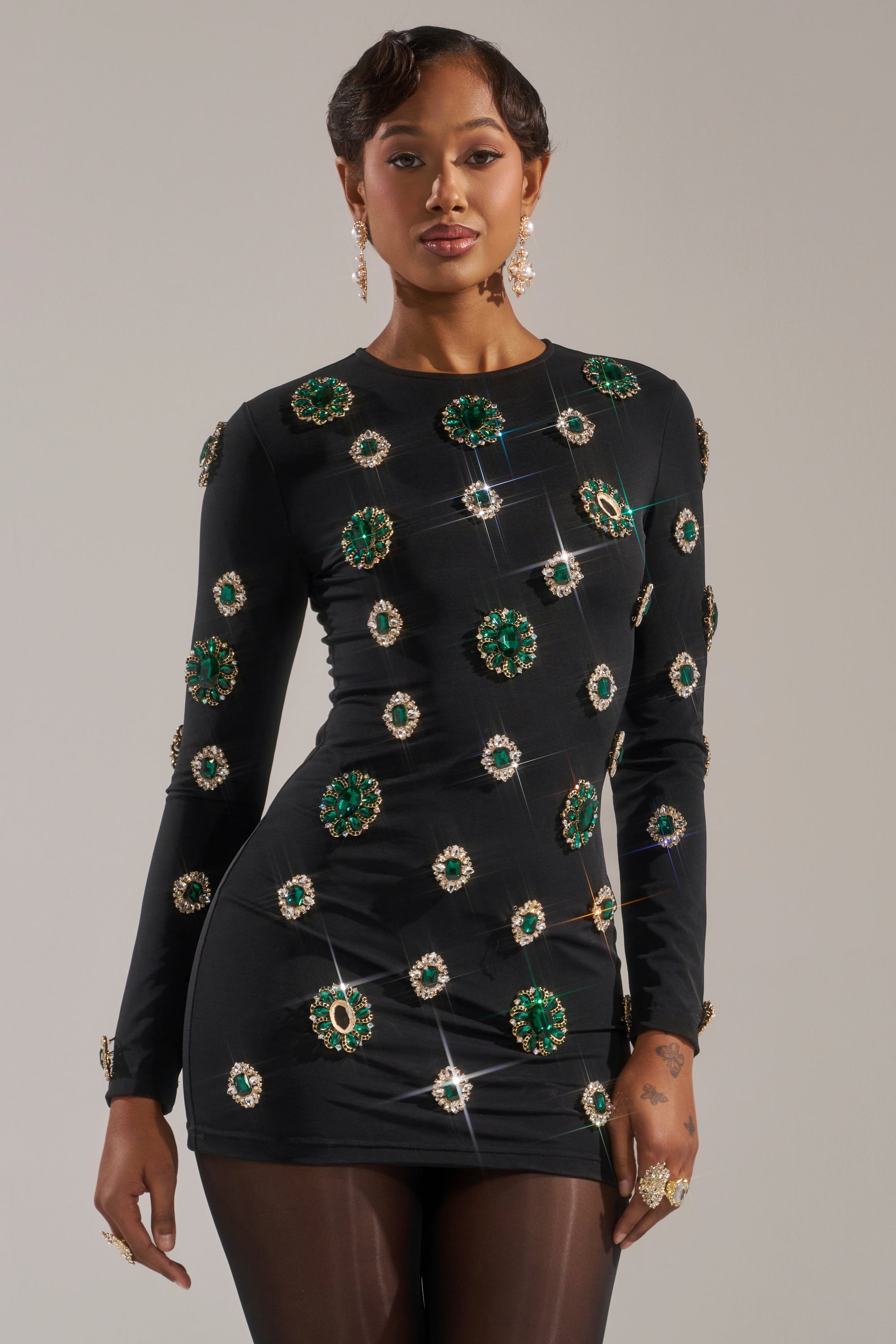 A woman models the BLING BLING MESH MINI DRESS WITH RHINESTONE EMBELLISHMENTS, a fitted black party dress adorned with sparkling green and gold rhinestones. She completes her glamorous look with earrings, rings, and soft waves in her hair.