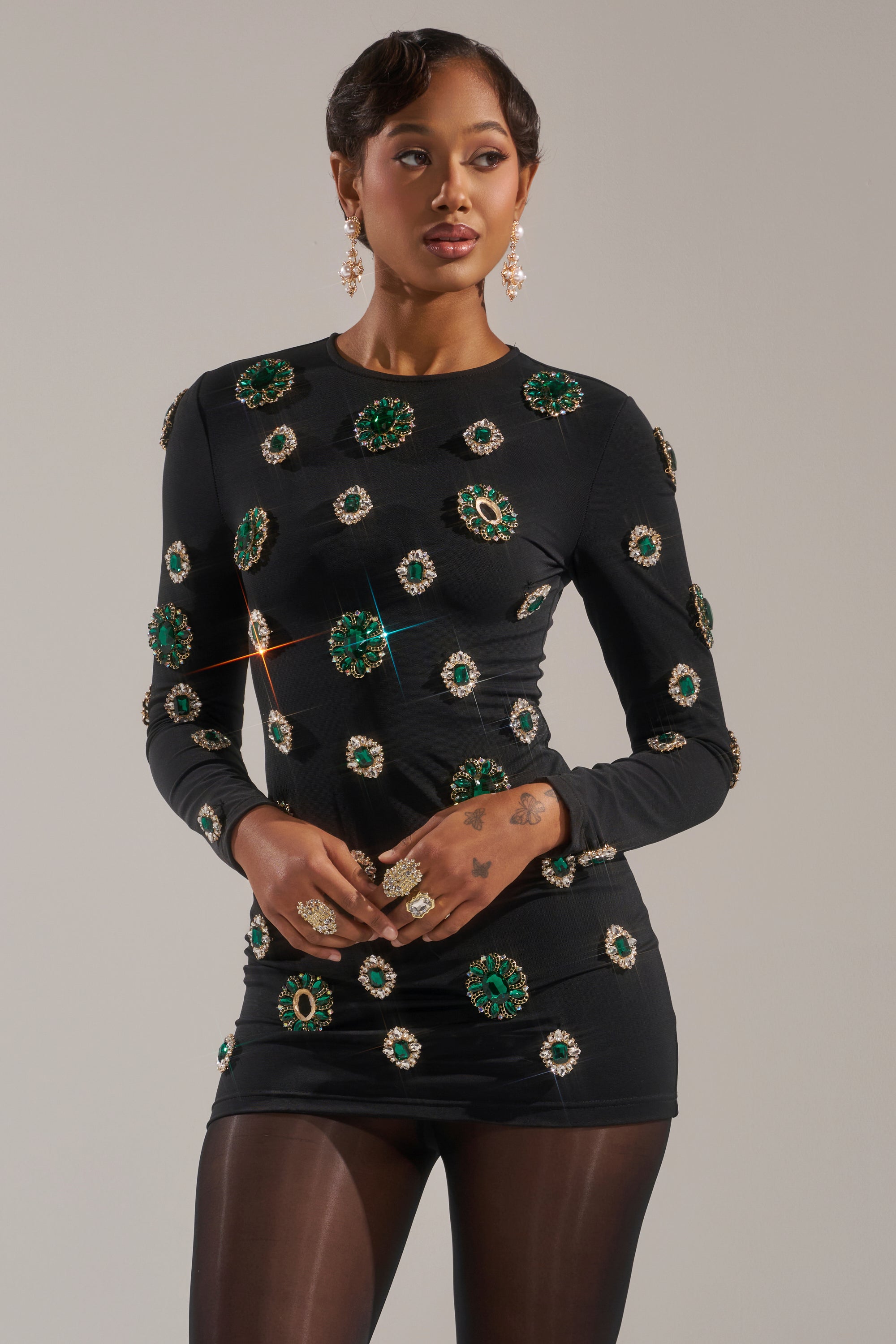 A woman poses against a plain light background in the BLING BLING MESH MINI DRESS WITH RHINESTONE EMBELLISHMENTS—an alluring, curve-hugging black long-sleeve mini dress featuring green and gold rhinestones. She completes the look with black tights, statement earrings, and rings.