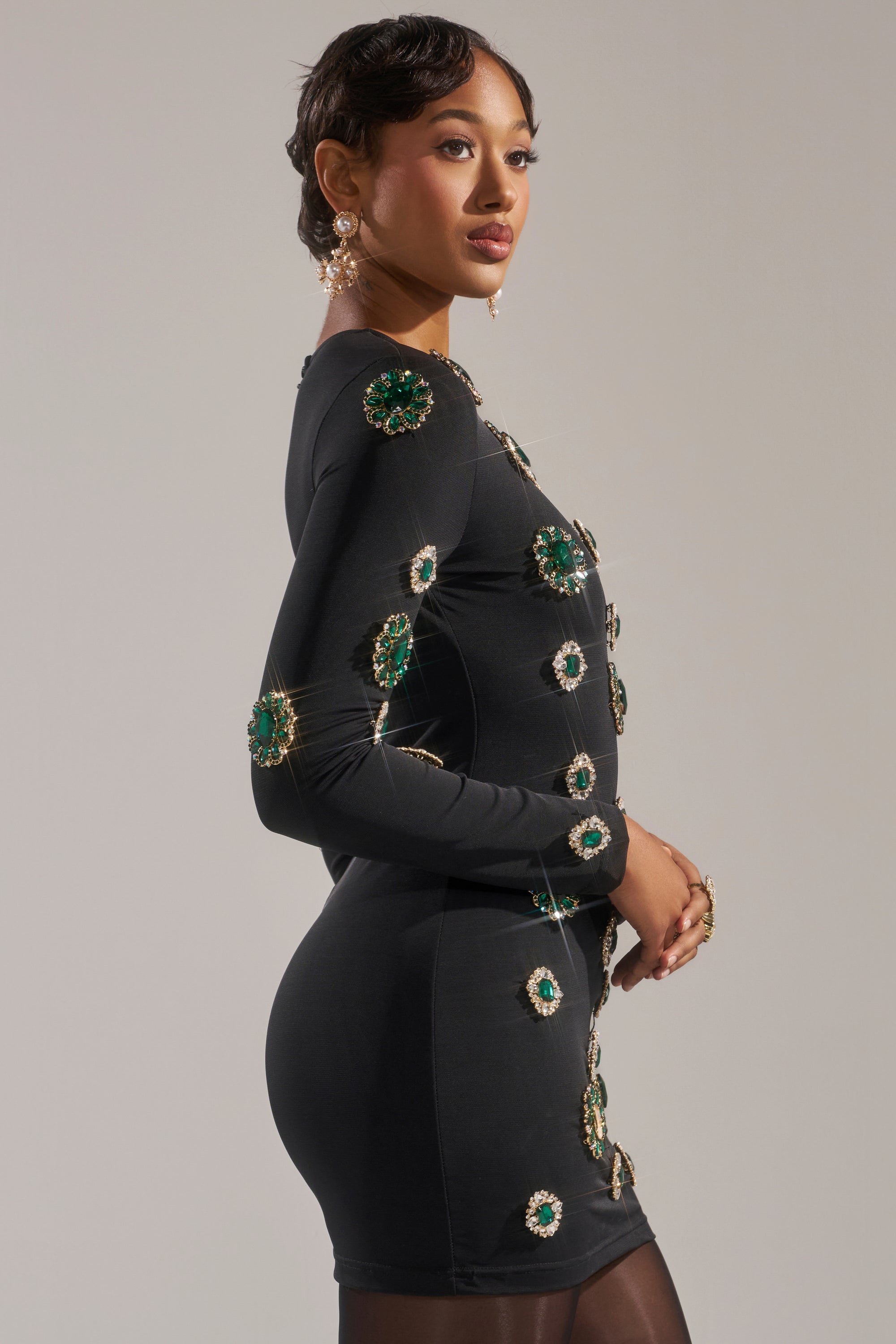 A person poses in the BLING BLING MESH MINI DRESS WITH RHINESTONE EMBELLISHMENTS— a fitted black dress featuring bold green and gold jeweled accents—styled with statement earrings and rings against a neutral background.