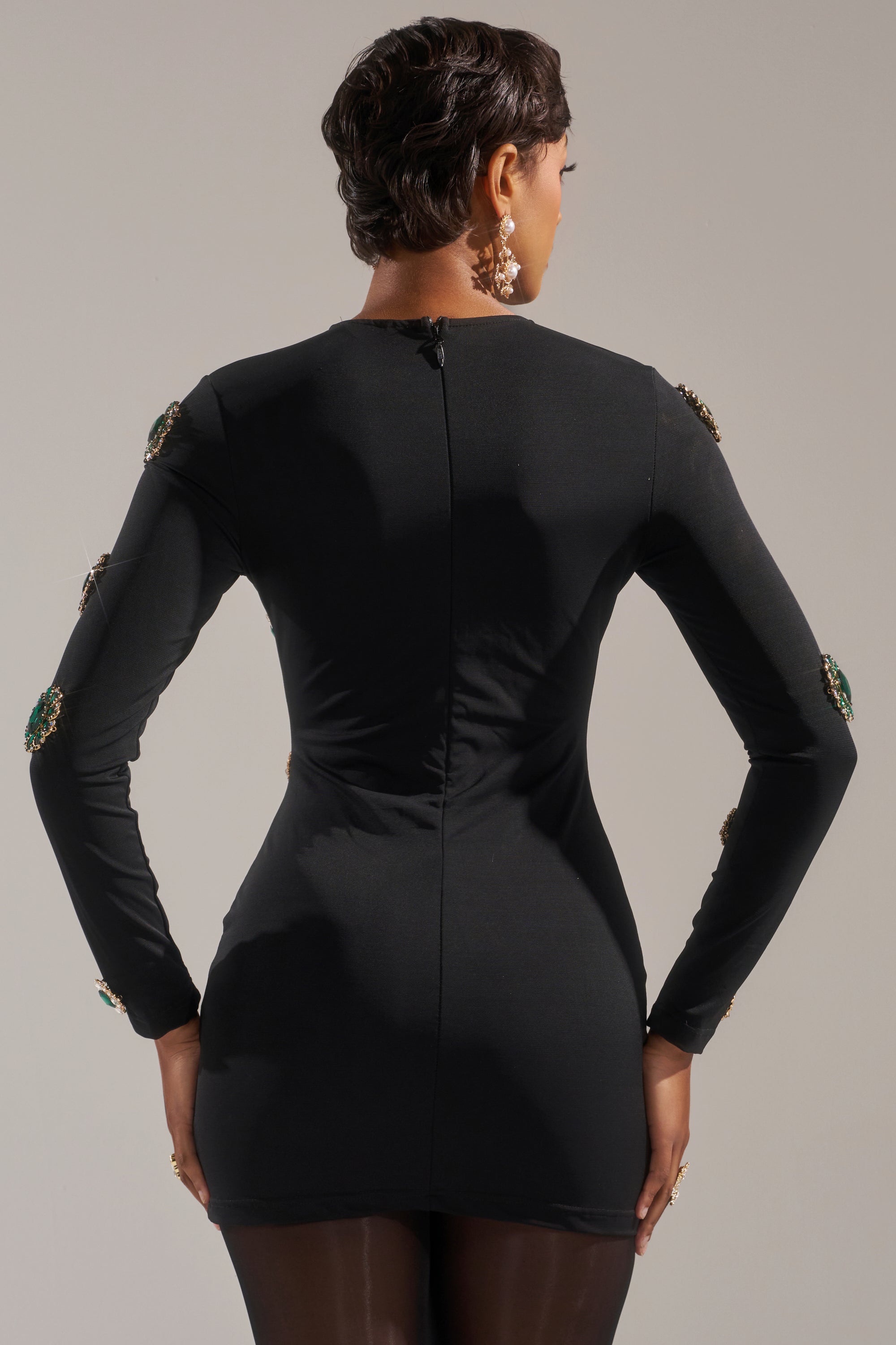 A woman with short hair models the BLING BLING MESH MINI DRESS WITH RHINESTONE EMBELLISHMENTS, a curve-hugging long-sleeve black dress featuring sparkling rhinestones on the sleeves. She accessorizes with bold, dangling earrings.