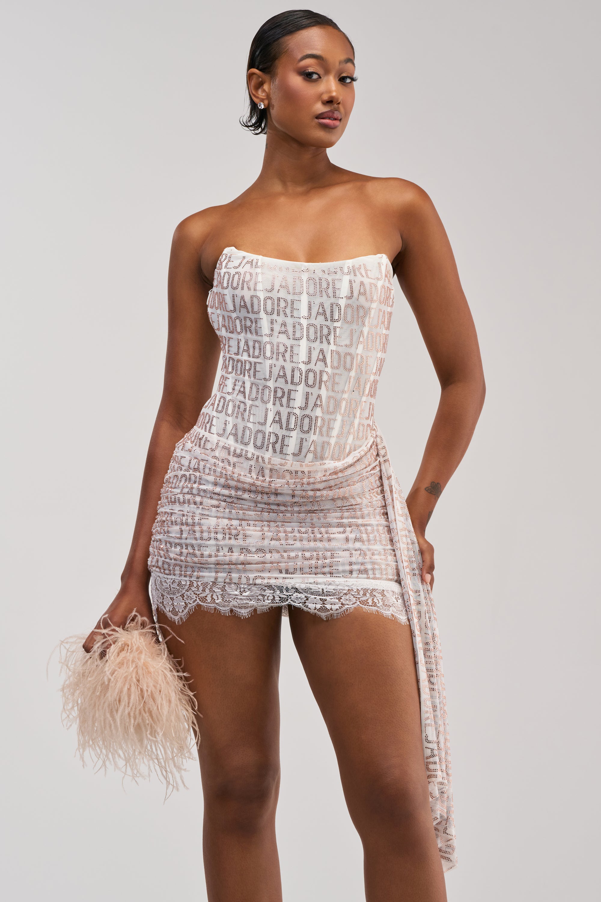A woman stands confidently in the LOVE ME LOVE ME RHINESTONE MINI DRESS IN WHITE, featuring a strapless cut and sparkling rhinestone details. She holds a feathery handbag, with her short, sleek hair and neutral expression set against a plain light background.