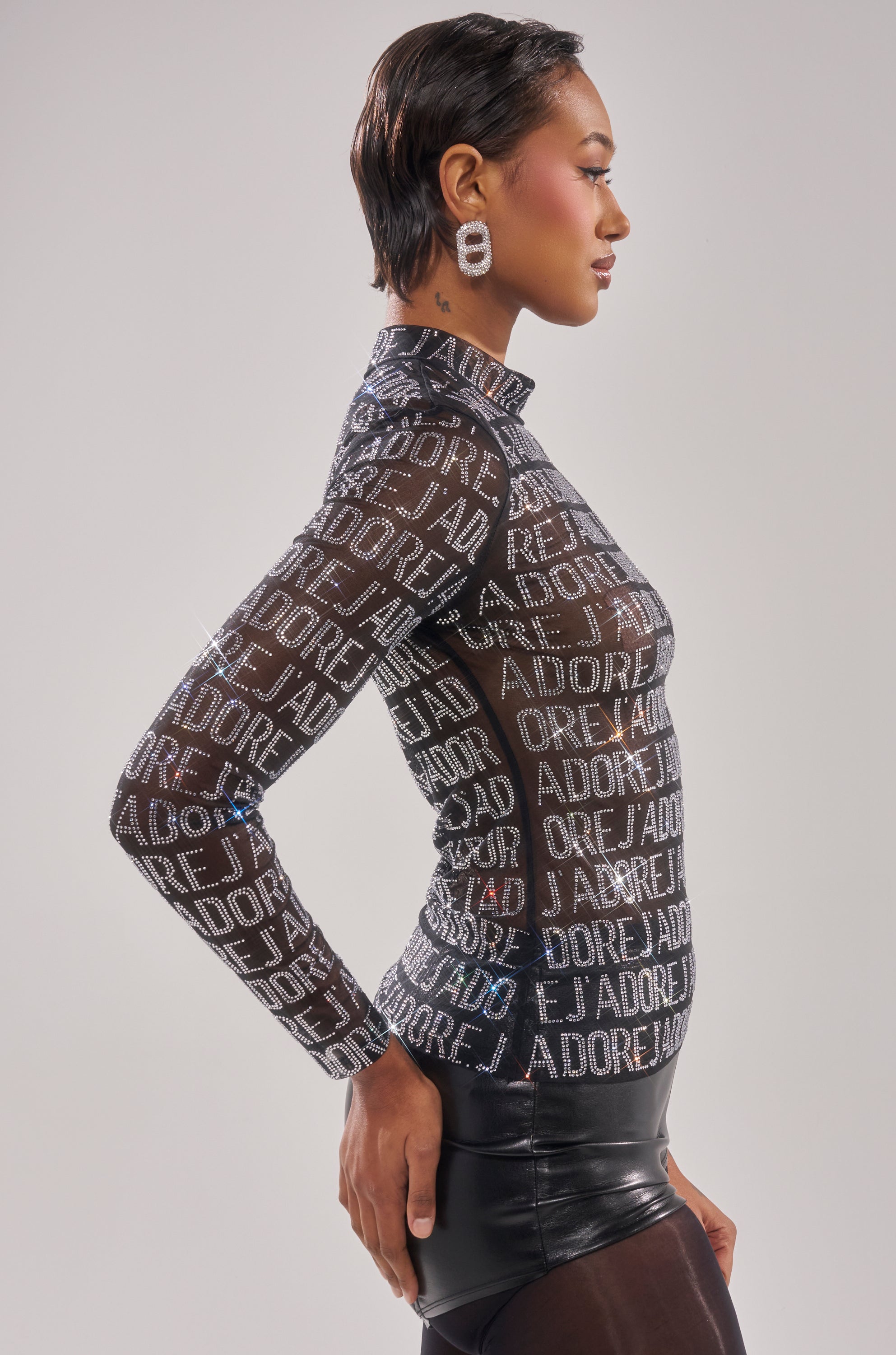 A woman with short hair poses in profile, wearing the J'ADORE EMBELLISHED MESH TOP featuring a bold "ADORE!" print, styled with black high-waisted pants and large rectangular earrings. 2