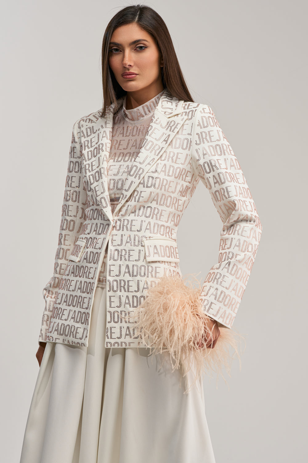 A woman with straight brown hair models the J'ADORE EMBELLISHED BLAZER IN WHITE, featuring light brown lettering, over a sheer beige top and white skirt. She accessorizes with a pale feathered clutch against a plain background.