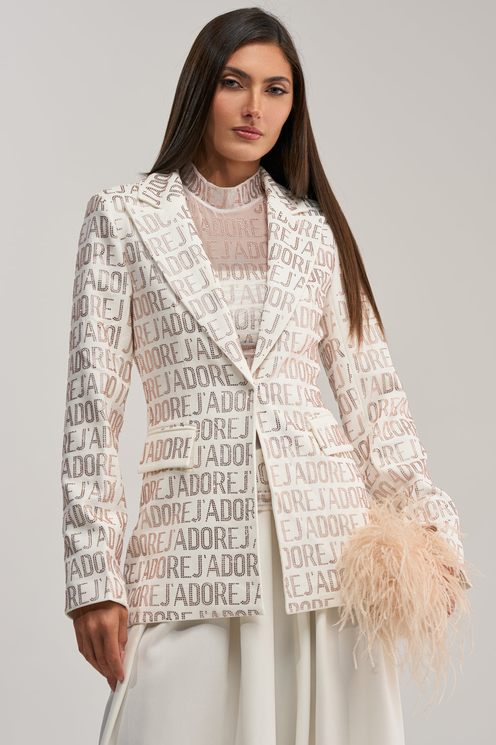 A woman with straight brown hair wears the J'ADORE EMBELLISHED BLAZER IN WHITE over a sheer high-neck blouse and white skirt, holding a feathery light beige accessory in one hand.