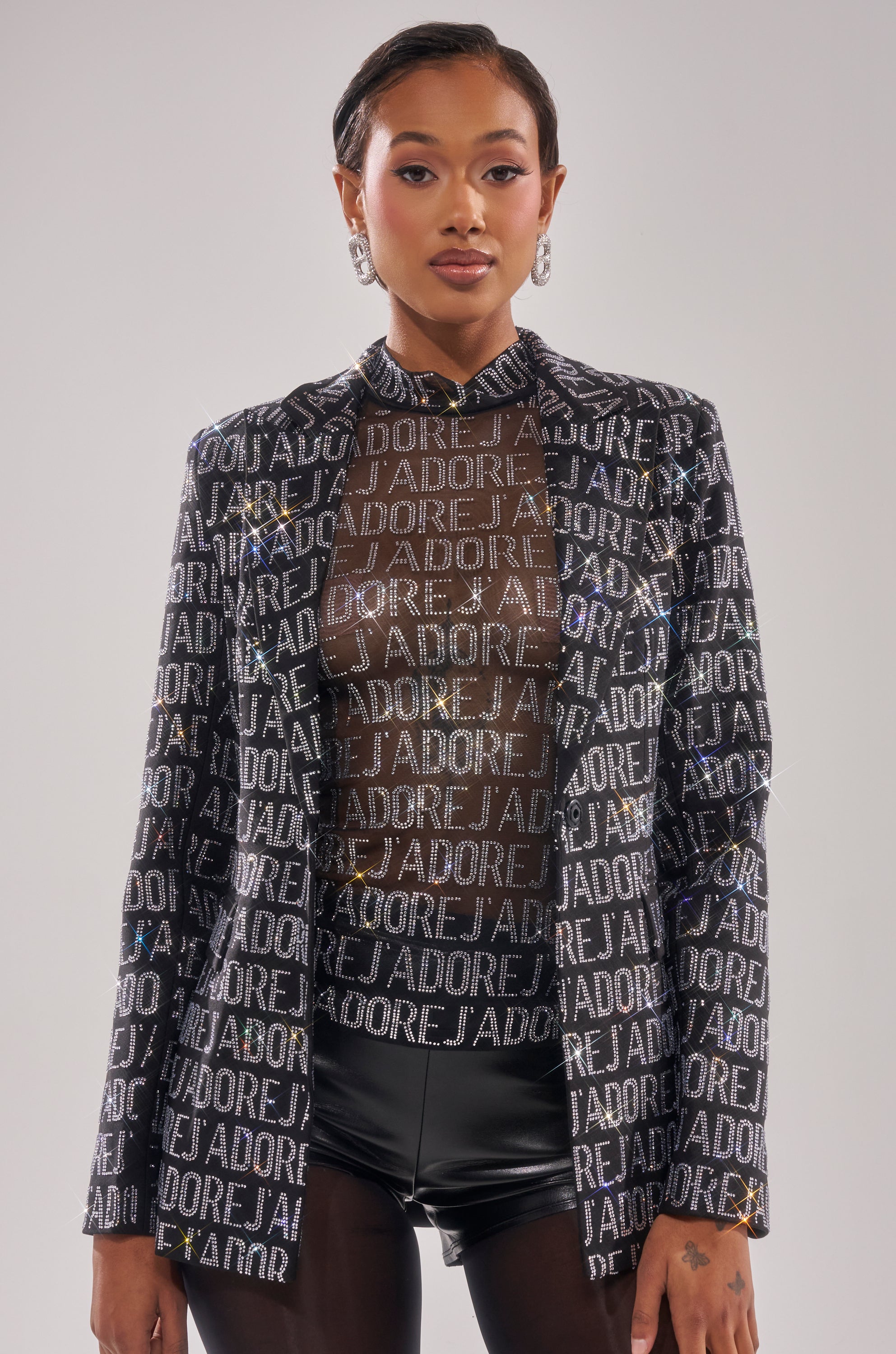 A person faces forward, wearing the J'ADORE EMBELLISHED BLAZER adorned with metallic letters, styled with a sheer top, black shorts, slicked-back short hair, and statement earrings.