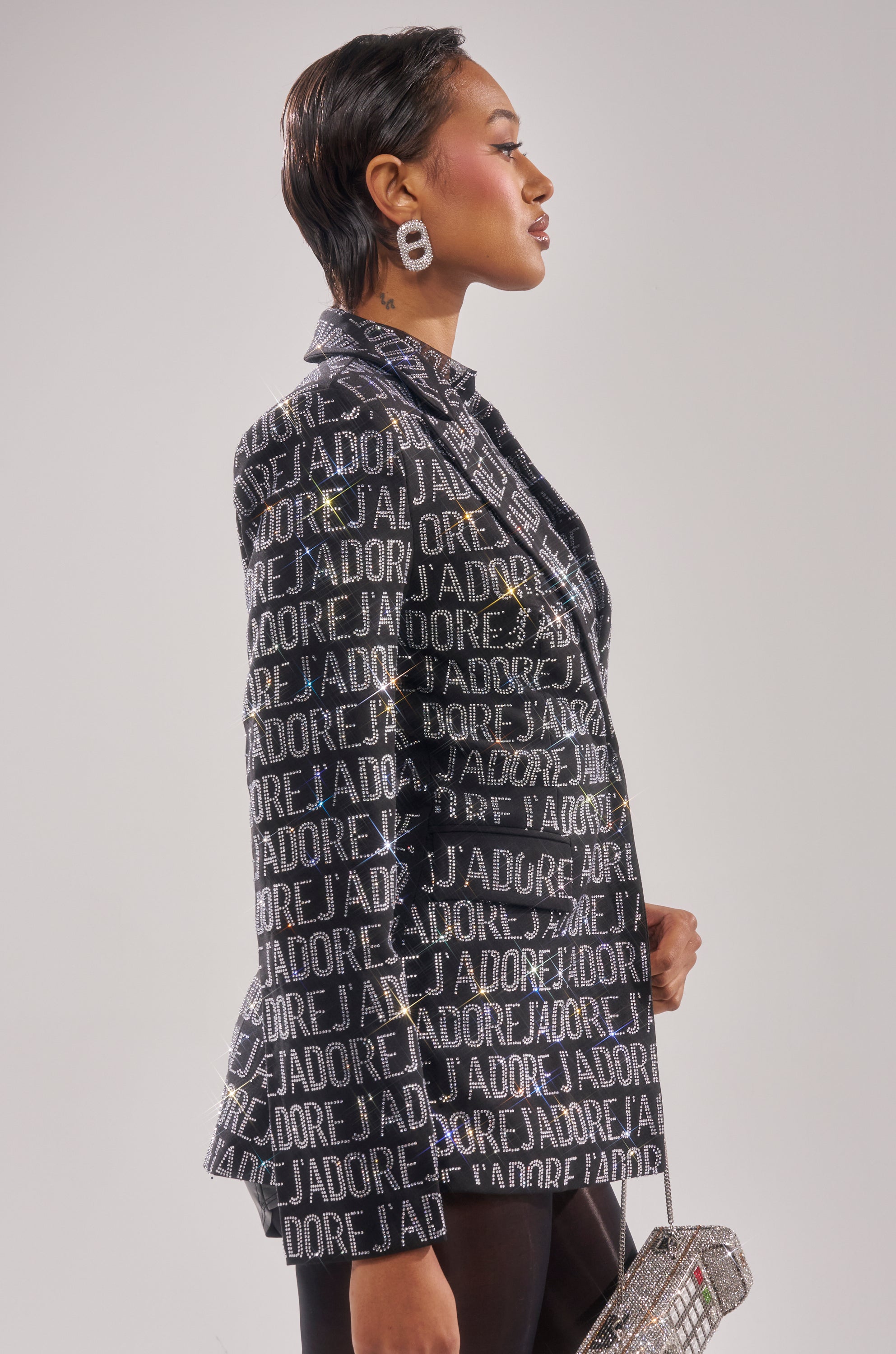 A person with short hair wears the J'ADORE EMBELLISHED BLAZER, styled with large geometric earrings and a glittery clutch, striking a pose against a plain background.