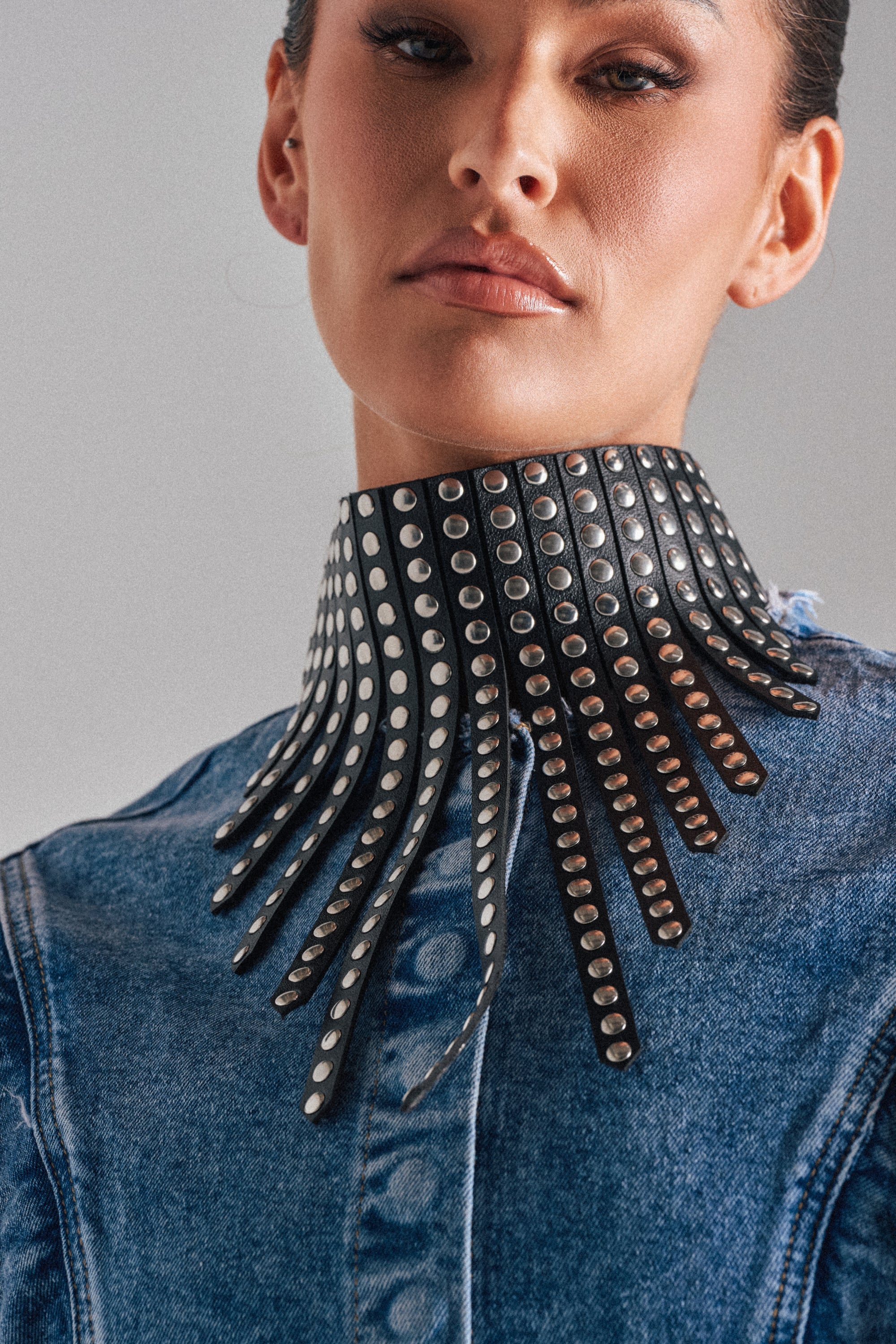 Someone in a denim shirt wears the EDGY VIBES NECKLACE, a bold faux leather choker featuring multiple fringed strips with silver studs that fan out around the neck. 0