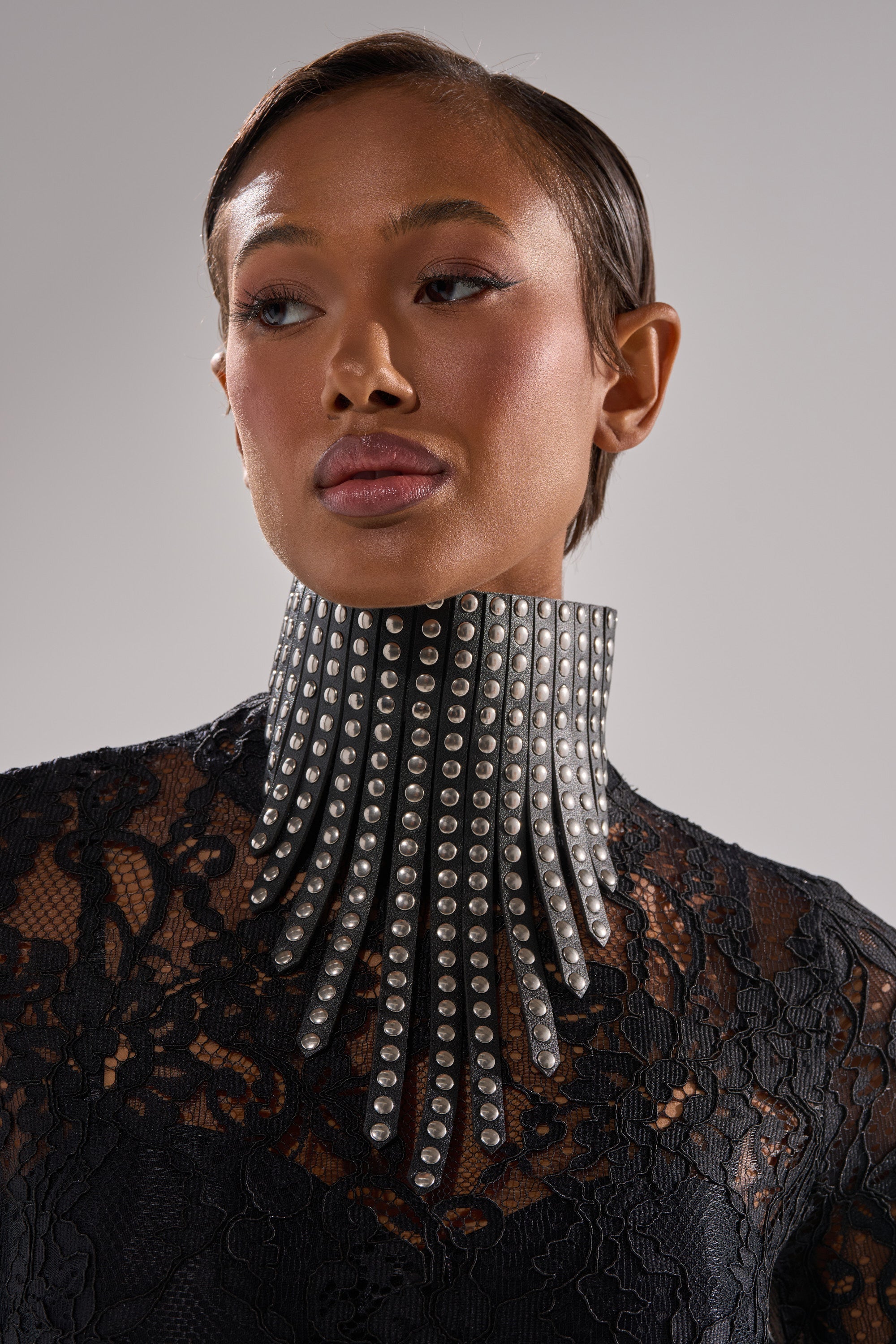 A person wears the EDGY VIBES NECKLACE—a bold black choker with silver studs and pointed extensions—paired with a black lace top against a plain background. 2