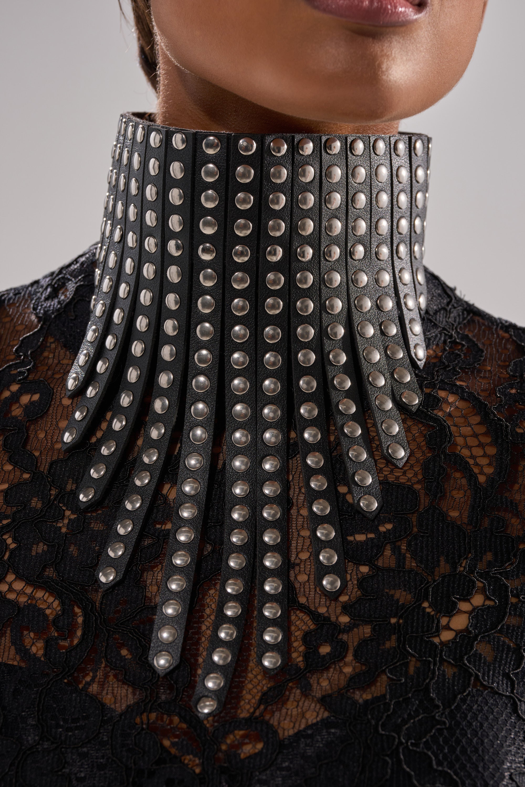 A person wears the EDGY VIBES NECKLACE—a dramatic, high black choker with wide, fanned-out vertical strips adorned with rows of silver studs—styled over a black lace top.