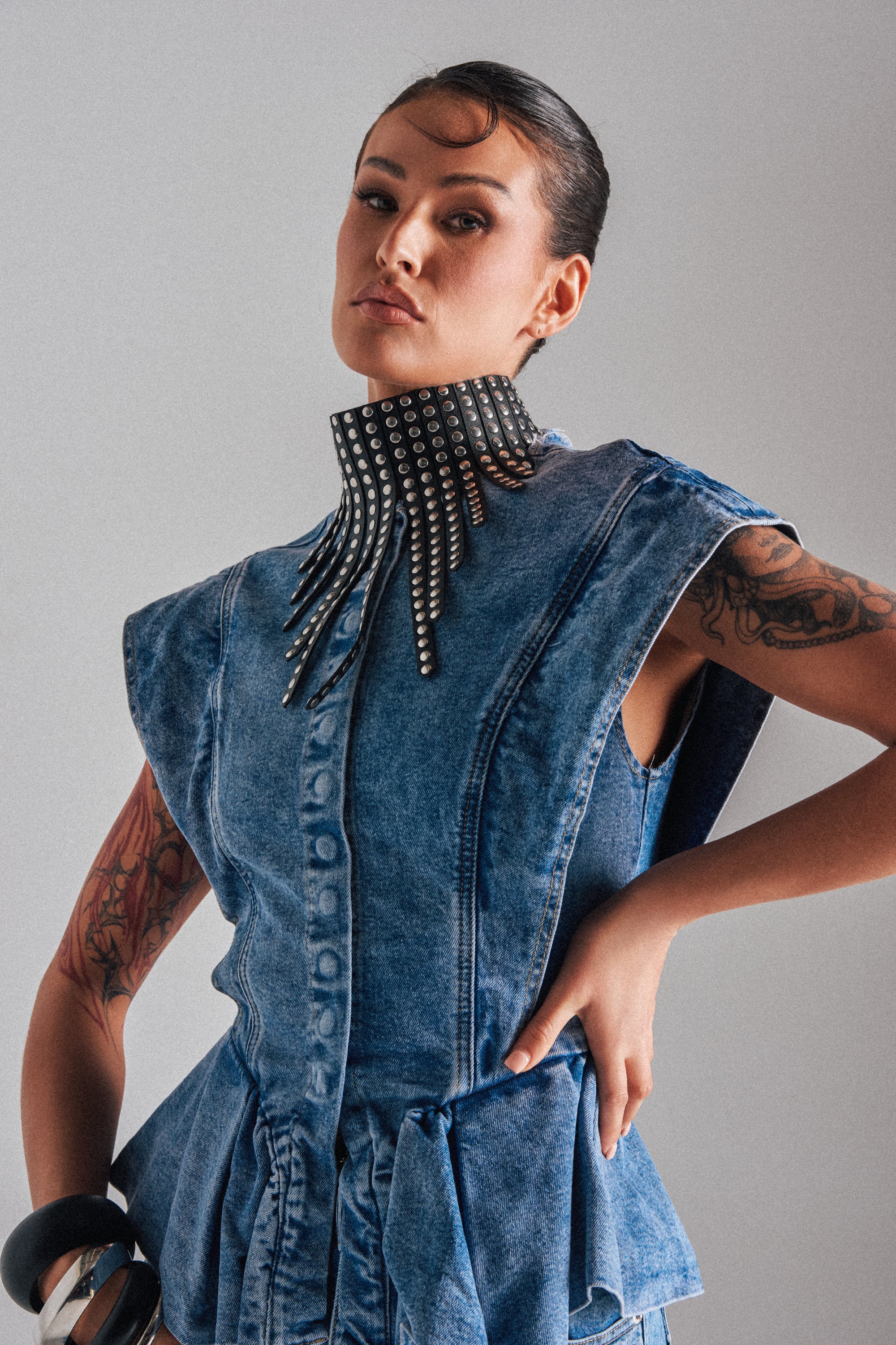 A tattooed woman exudes confidence in a sleeveless denim dress with structured shoulders, bold studded collar, chunky bangles, and the EDGY VIBES NECKLACE.