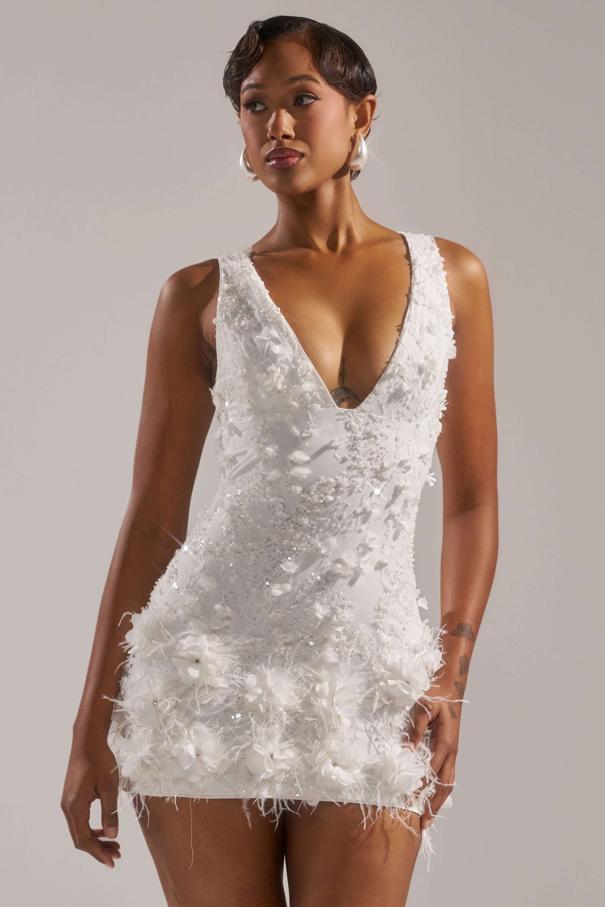 A woman wearing the WINTER KISS MINI DRESS, a sleeveless white mini dress with a deep V-neckline, floral textures, and feathered hem, stands against a plain background.