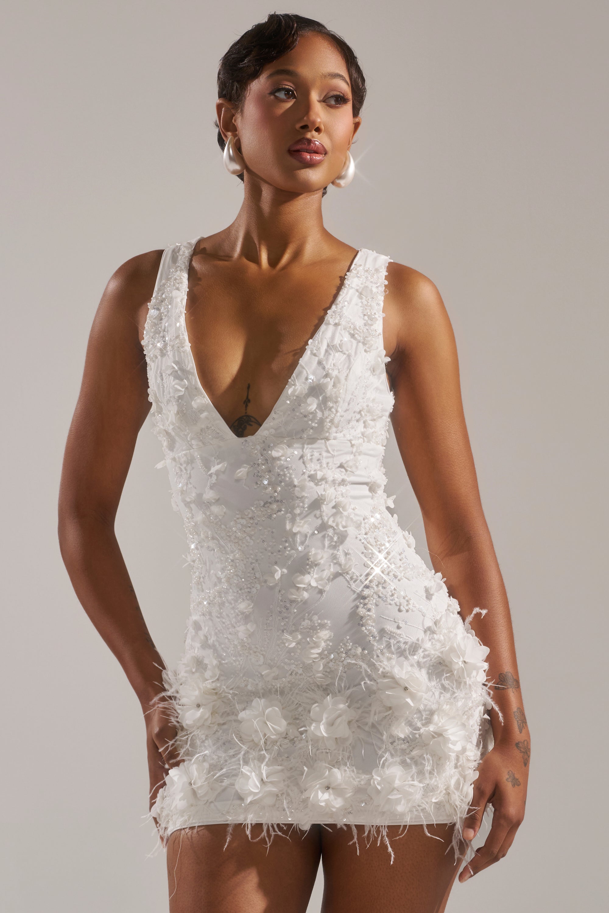 A woman in the WINTER KISS MINI DRESS—a sleeveless, white V-neck mini dress with floral appliqué and textured details—stands against a plain background.