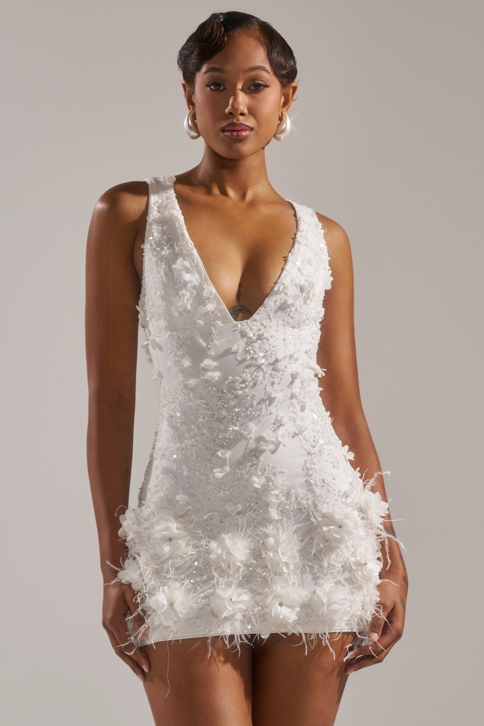 A woman stands facing forward against a plain, light background, wearing the WINTER KISS MINI DRESS—a white sleeveless V-neck mini dress decorated with floral appliqués and finished with a feather hem.
