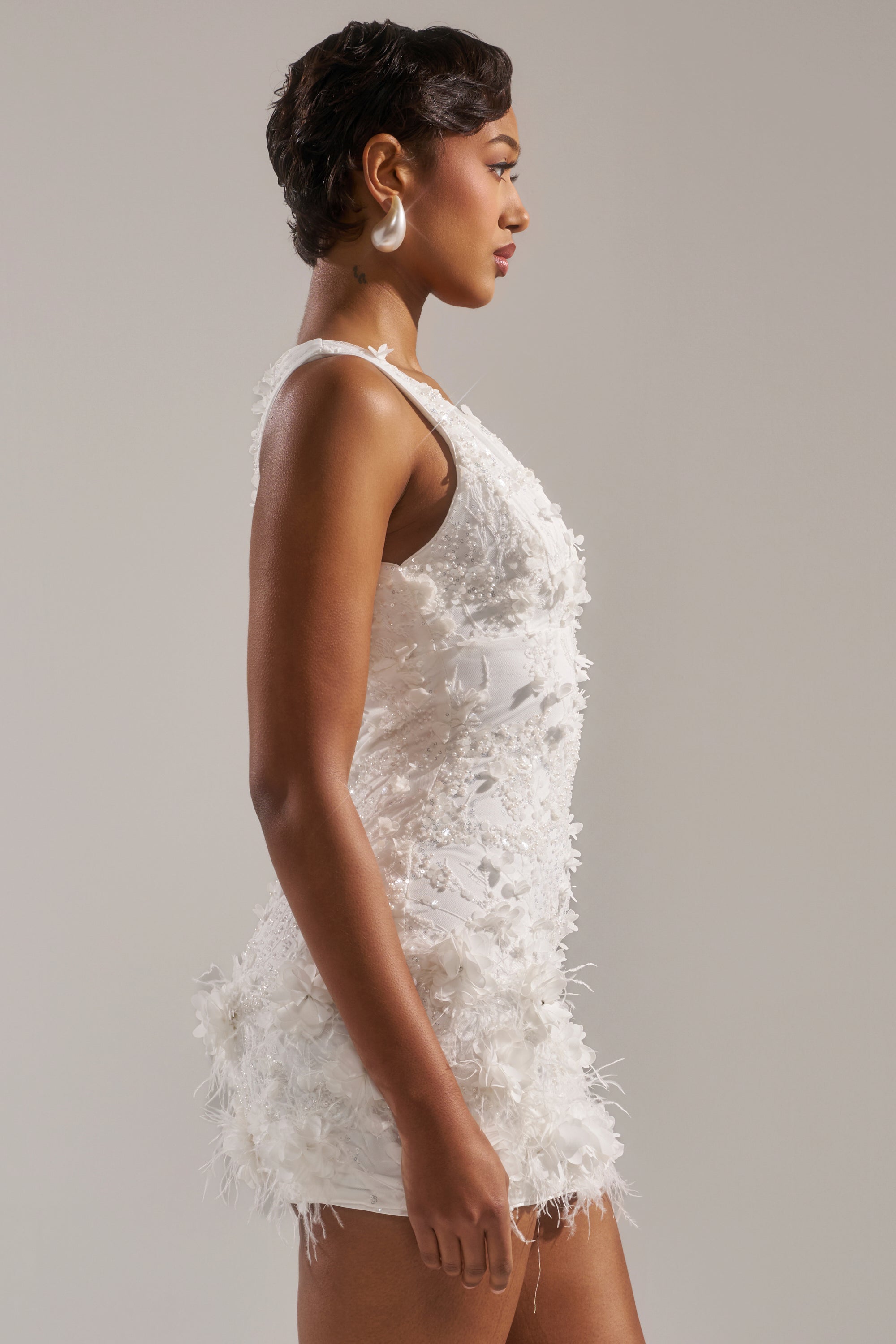 A woman with short, styled hair models the WINTER KISS MINI DRESS—a sleeveless white mini dress with a feathered hem and floral accents—while standing in profile against a plain light gray background.
