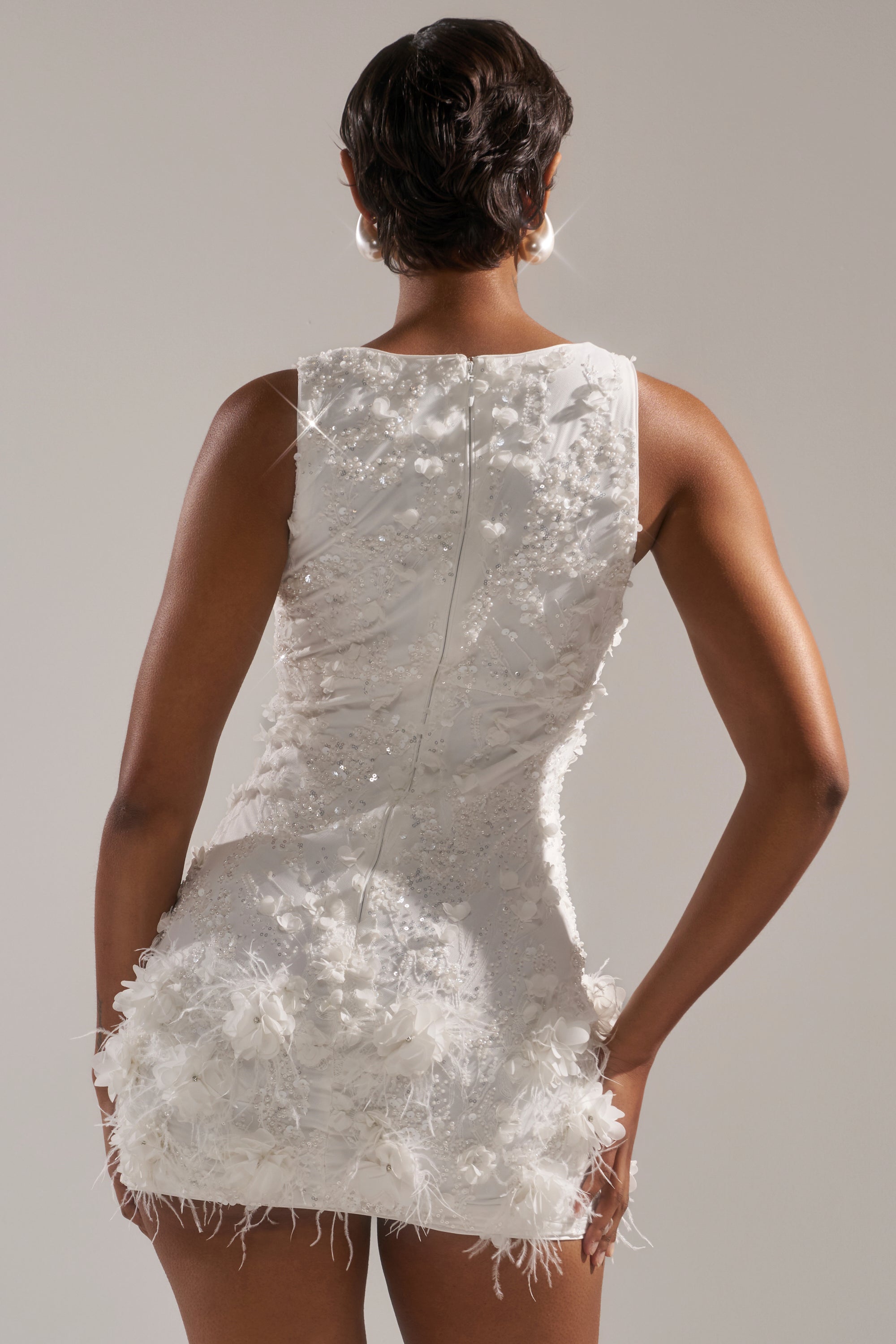 A woman with short hair stands against a light background wearing the WINTER KISS MINI DRESS, a sleeveless white dress featuring a feather hem and textured floral details.