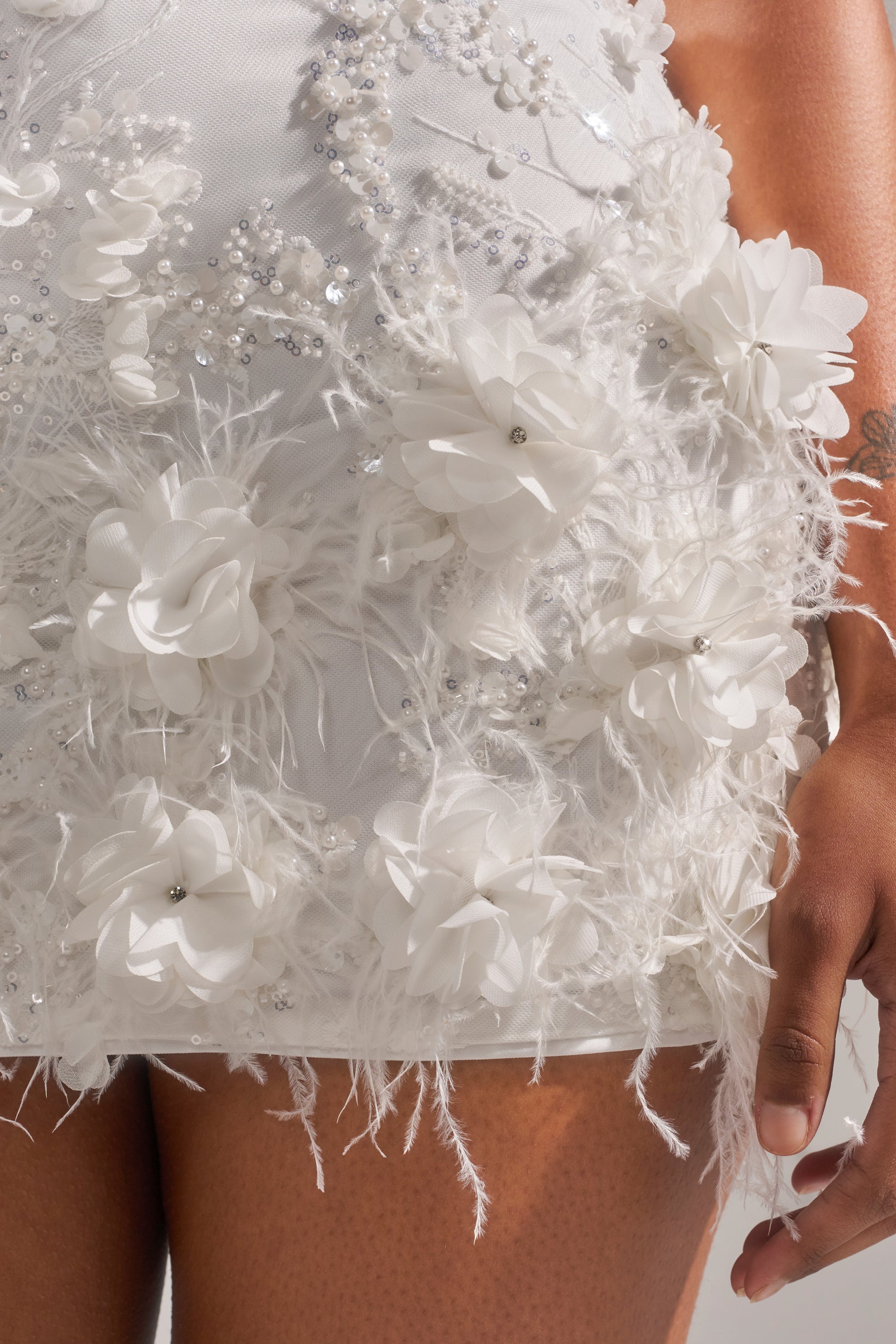 A close-up of someone in the WINTER KISS MINI DRESS, featuring 3D floral appliqués, beads, sequins, and a feather hem. One hand by their side reveals a small arm tattoo.