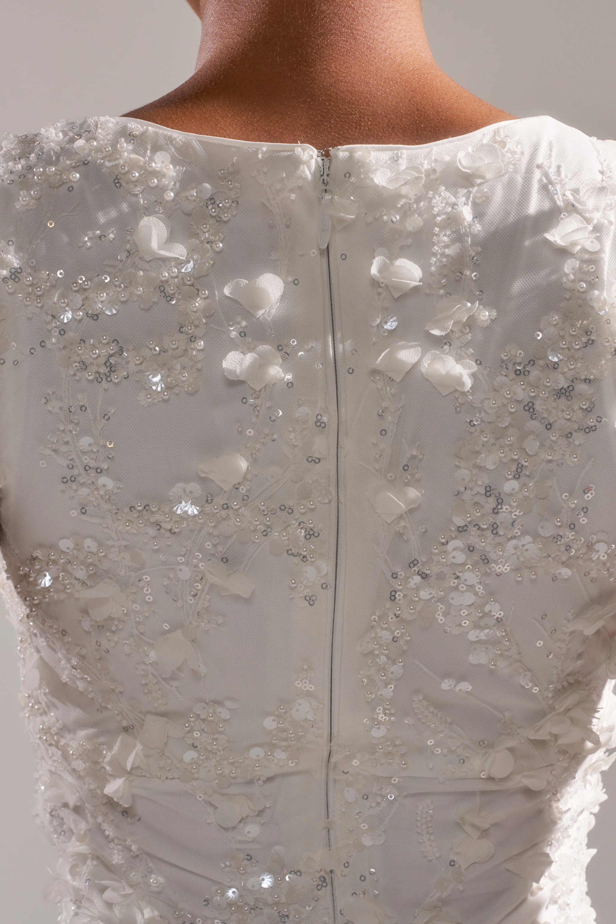 Close-up of the WINTER KISS MINI DRESS: a white sequin mini dress with intricate floral embroidery, beads, and sequins. Features a visible center zipper and an open back revealing the upper back and neck.