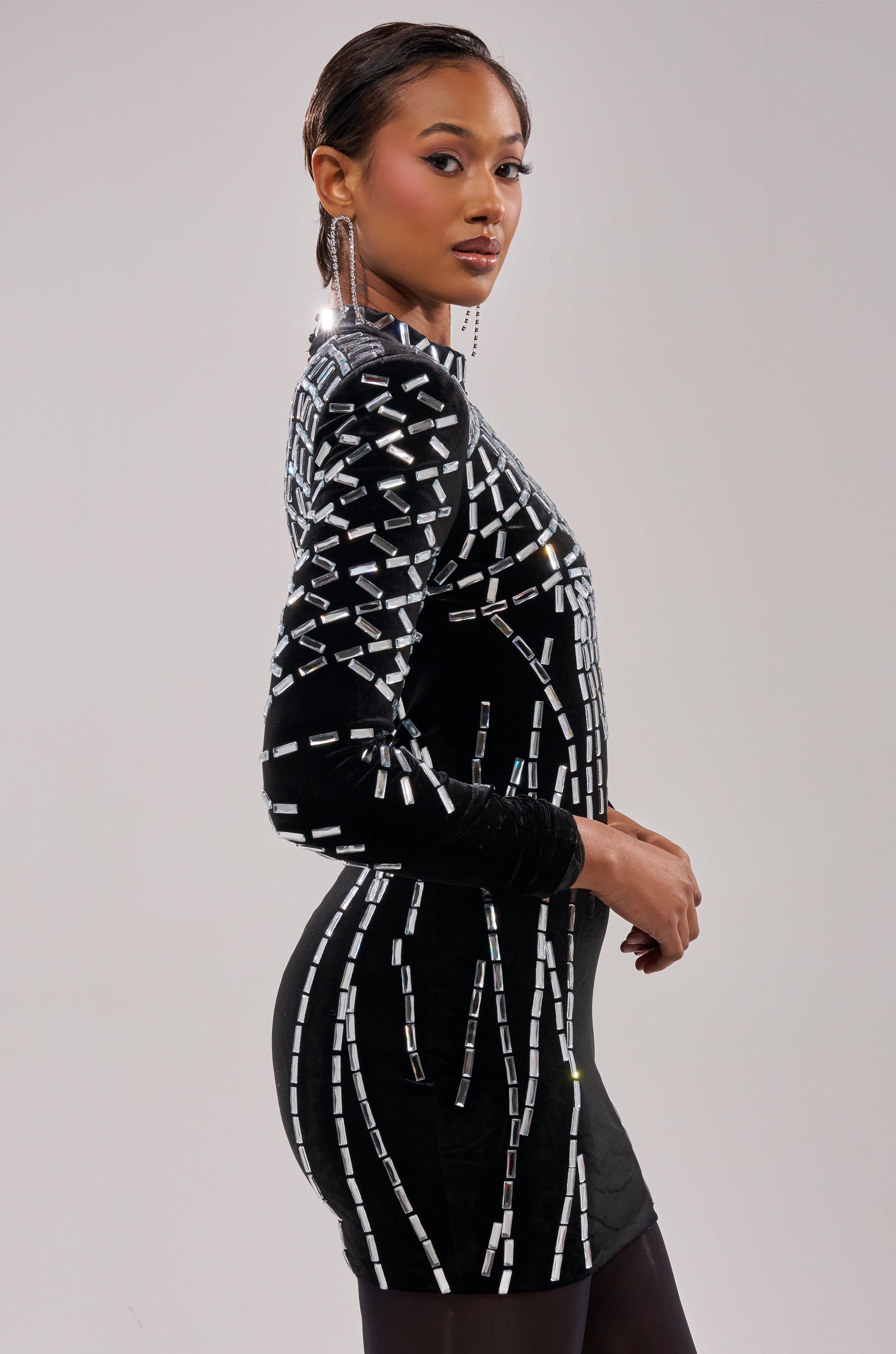 A woman stands in profile against a plain background, wearing the FIRECRACKER RHINESTONE MINI DRESS—a fitted black mini dress with silver geometric patterns. She has sleek, side-parted hair, statement earrings, and a confident expression.