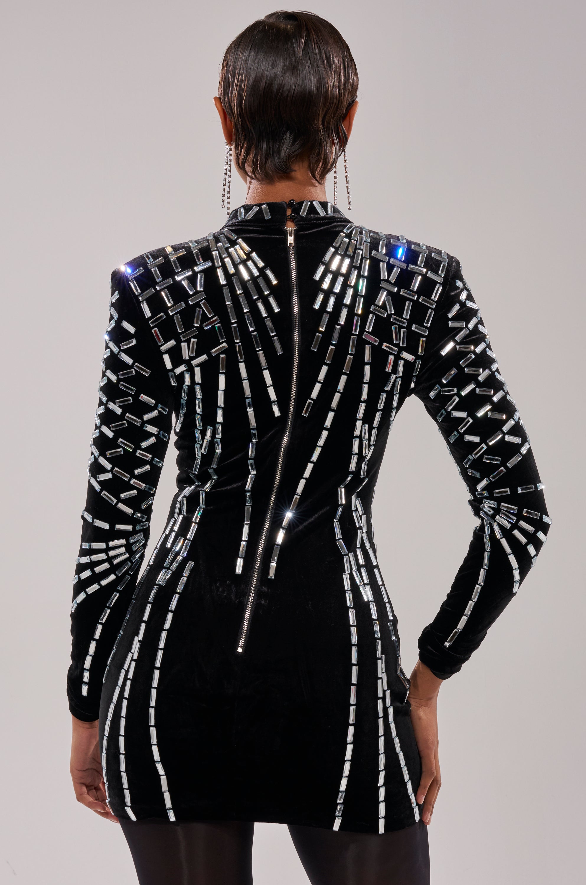 A person faces away from the camera, modeling the FIRECRACKER RHINESTONE MINI DRESS—a fitted black mini dress with reflective rhinestone embellishments and a visible back zipper, set against a plain light backdrop.