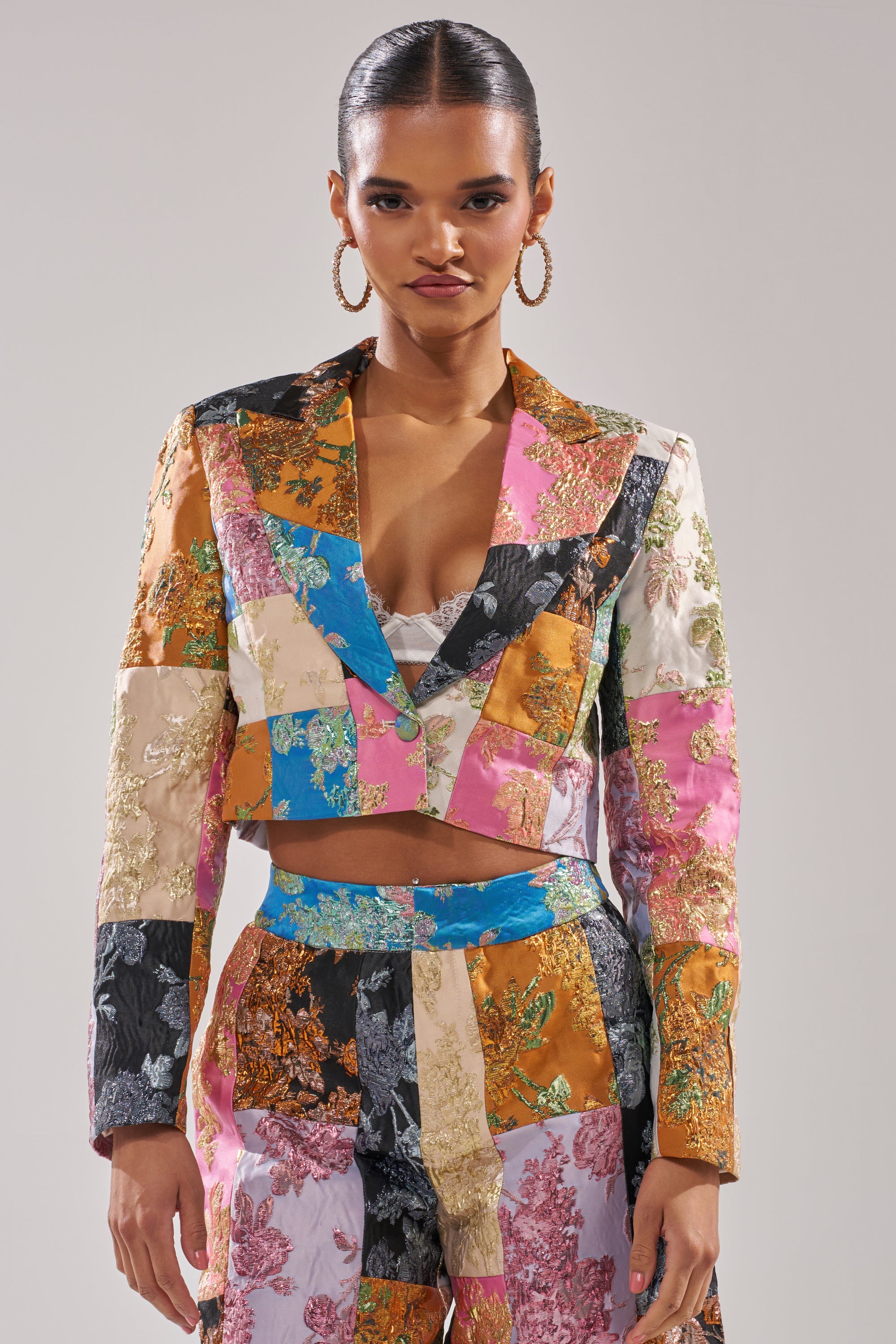 A woman exudes confidence in the LOLITA PATCHWORK BROCADE BLAZER with intricate floral patterns and matching pants. Her slicked-back hair and bold hoop earrings complete the look, set against a plain background. 0