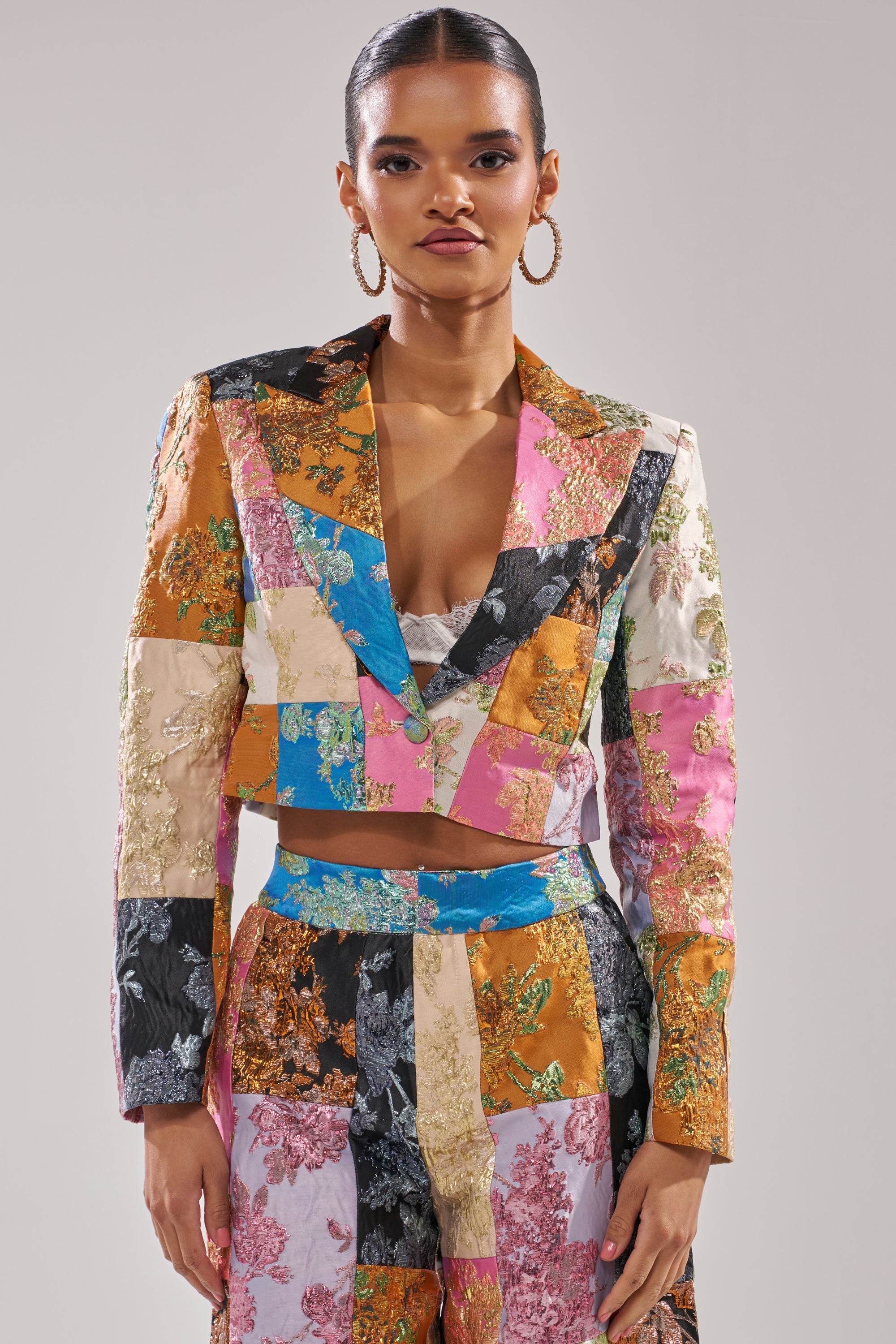 A woman stands confidently against a plain background wearing the LOLITA PATCHWORK BROCADE BLAZER with embroidered accents and high-waisted pants. She accessorizes with hoop earrings and has her hair pulled back. 1