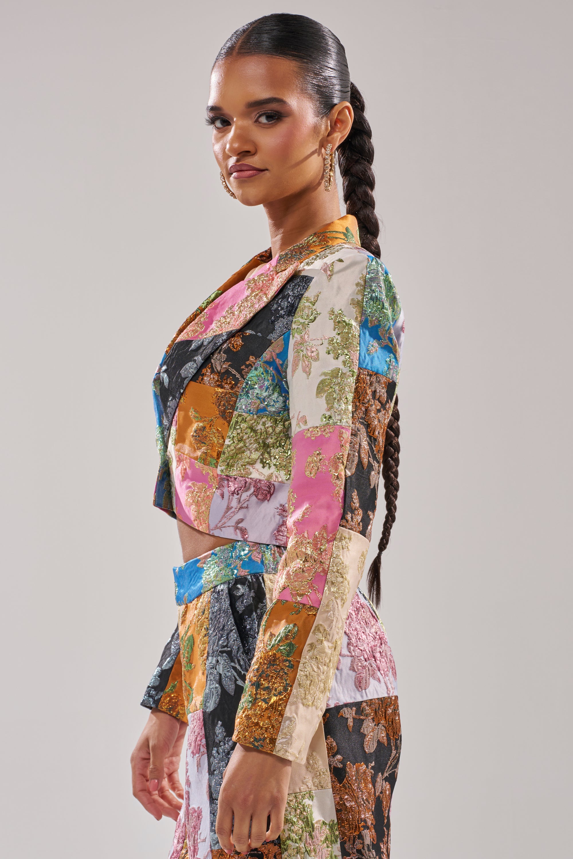 A woman with long, dark, braided hair stands in profile, wearing the LOLITA PATCHWORK BROCADE BLAZER with colorful floral patterns. The background is plain and light-colored. 2