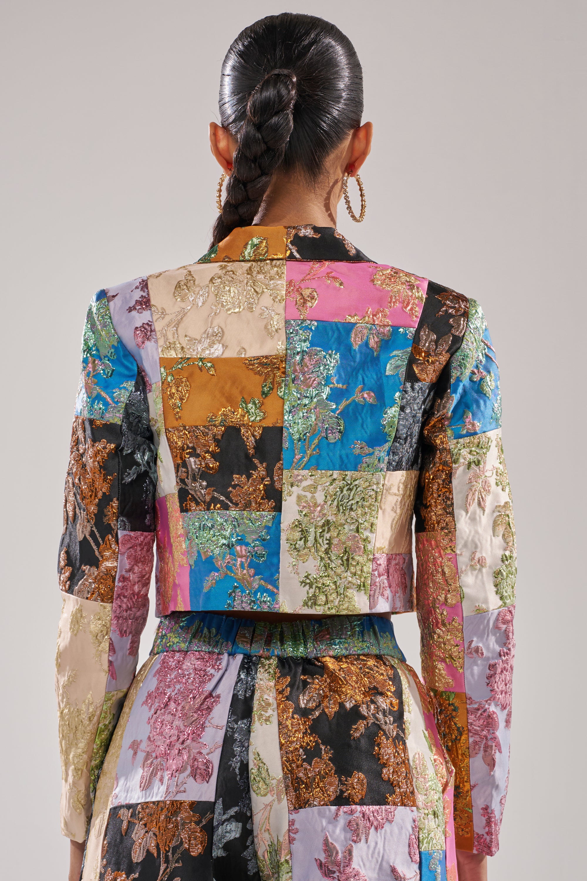 A woman with a long, braided ponytail and large hoop earrings is seen from behind wearing the LOLITA PATCHWORK BROCADE BLAZER and matching pants with embroidered floral patterns, set against a plain background. 3