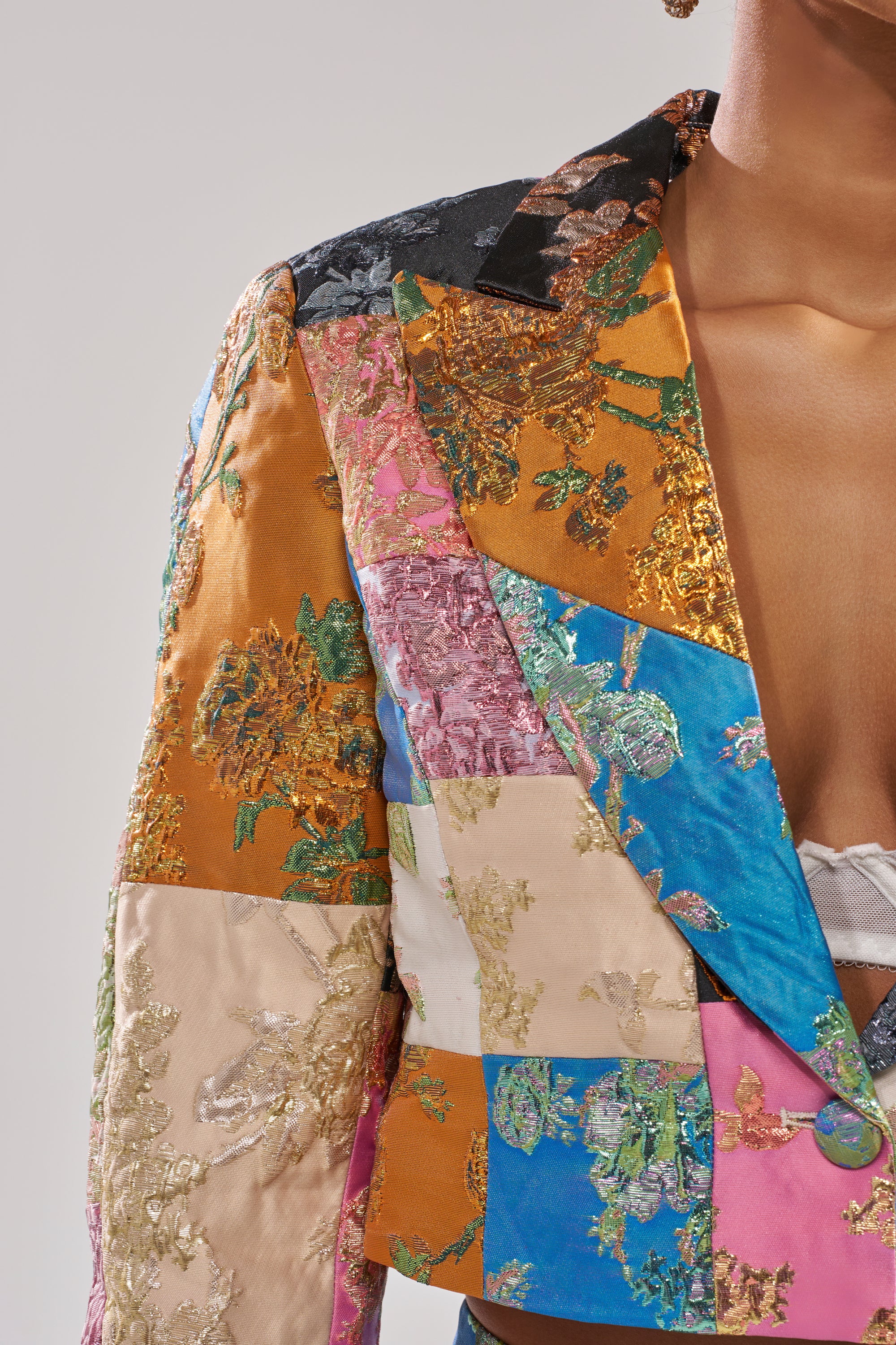 A person wears the LOLITA PATCHWORK BROCADE BLAZER—shiny, embroidered in gold, pink, blue, and orange—over a white bralette, shown from shoulder to waist against a plain background. 6